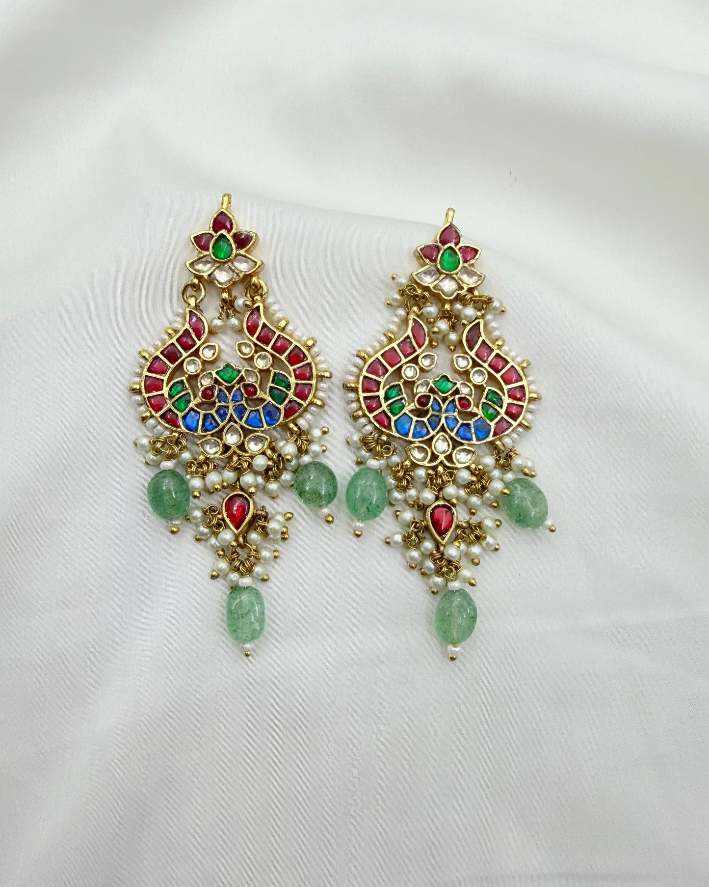 A pair of ornate gold-toned Indian-style chandelier earrings decorated with multicolored enamel, green stones, and small pearl-like beads, displayed on a white fabric background.
