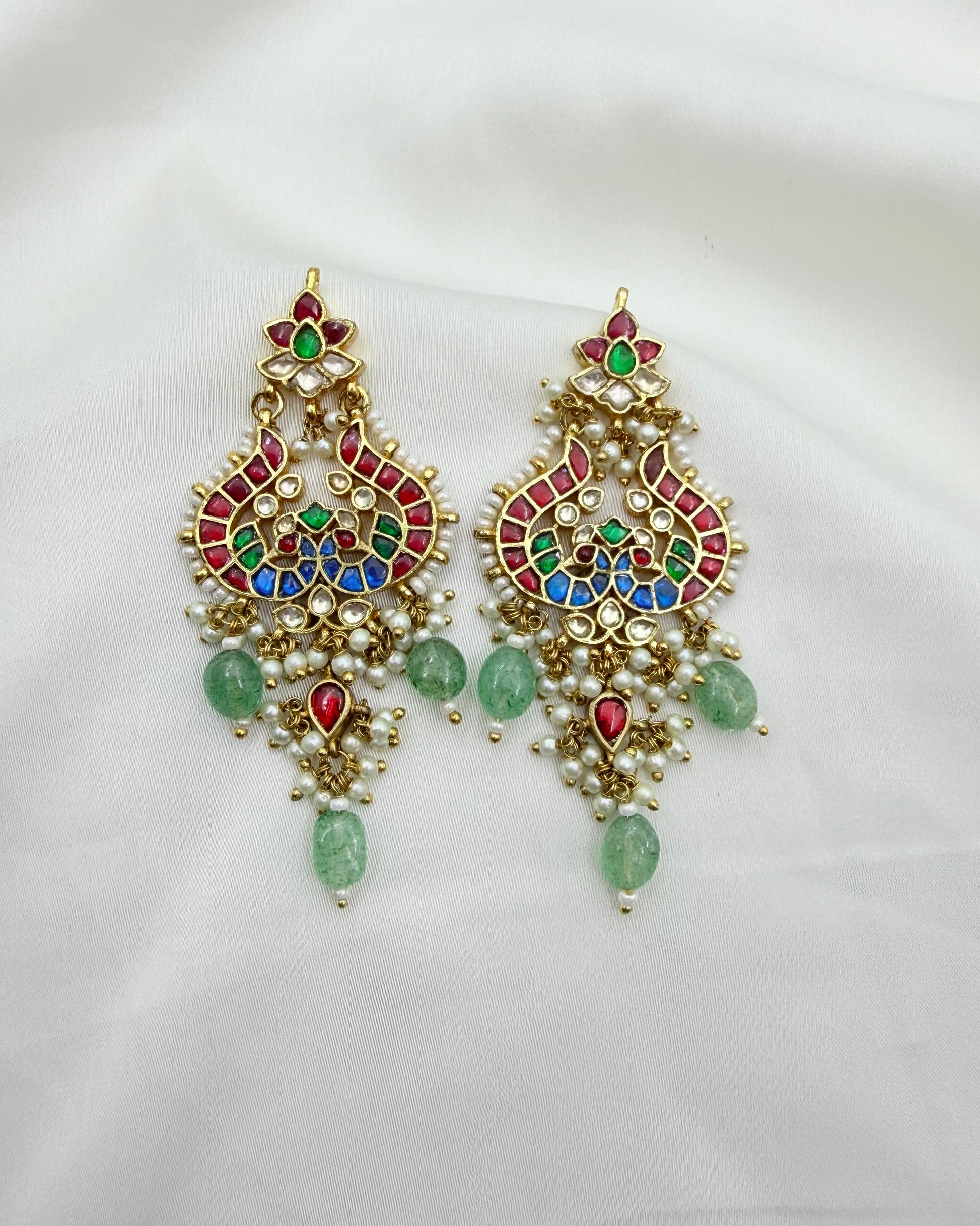 A pair of ornate gold-toned Indian-style chandelier earrings decorated with multicolored enamel, green stones, and small pearl-like beads, displayed on a white fabric background.