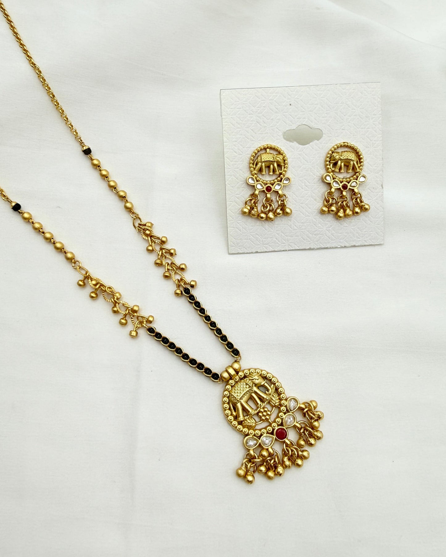 Gold-toned traditional jewelry set featuring a necklace with a detailed pendant and matching stud earrings, displayed on a white fabric background with the earrings on a small card.
