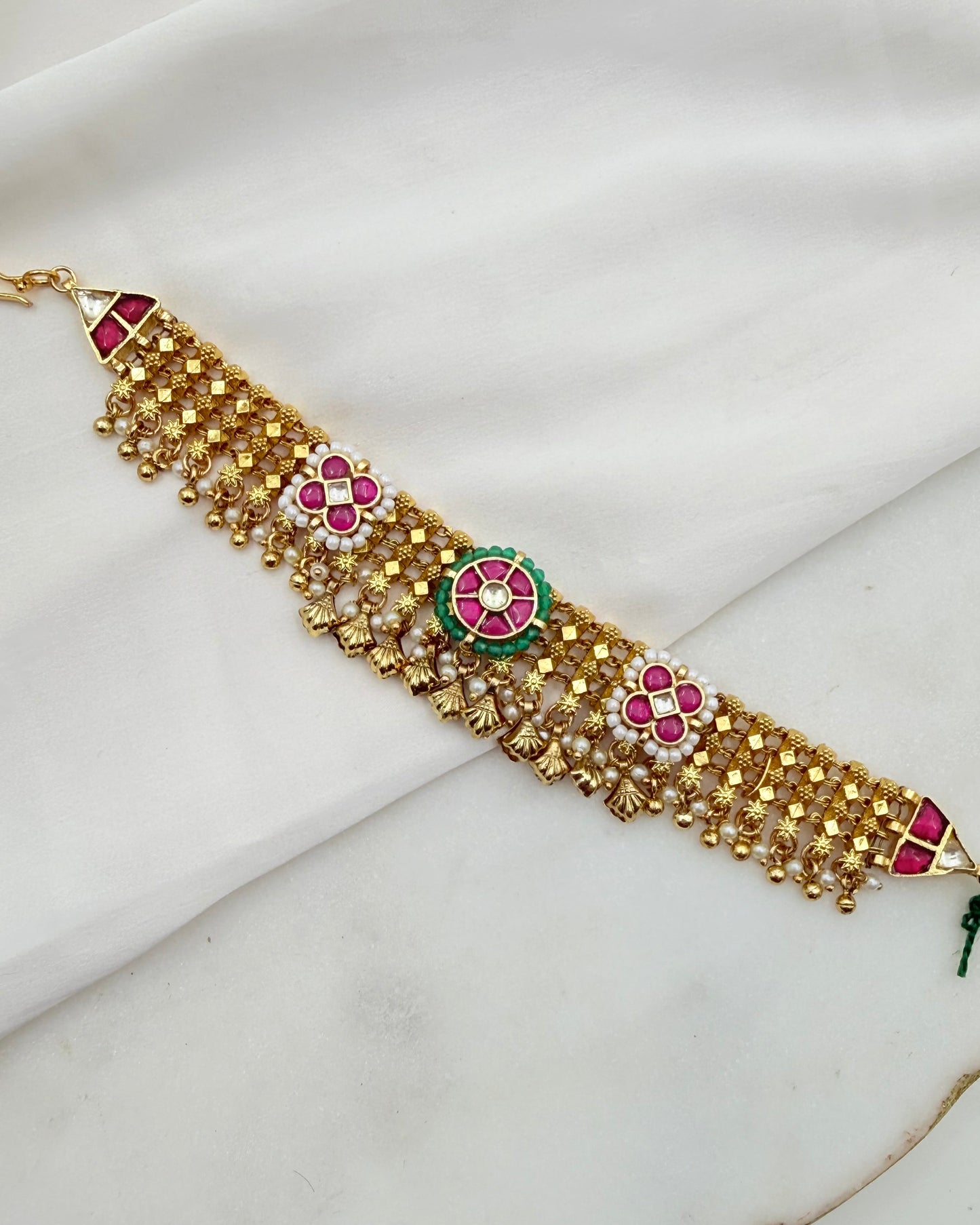 Gold-toned beaded bracelet with pink and green floral motifs and white stones, laid diagonally on a white fabric background.