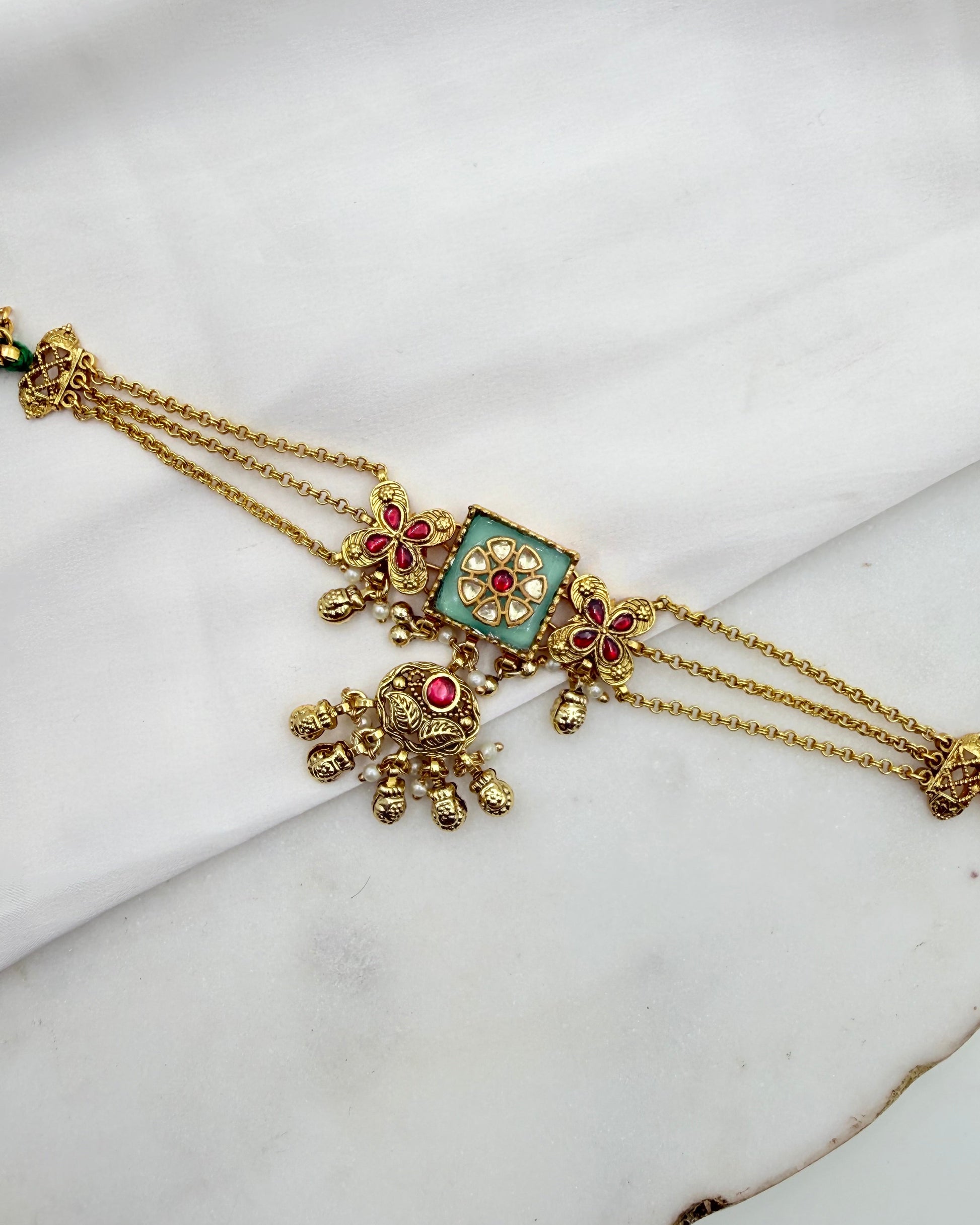 An ornate gold-toned traditional Indian jewelry piece, likely a choker or necklace, featuring a central square turquoise enamel panel with red floral accents and dangling gold filigree, displayed on a white fabric background.