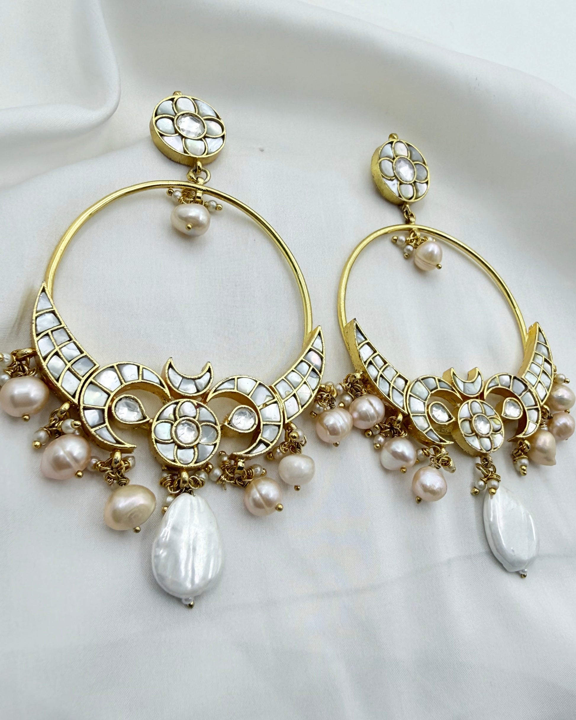 A pair of gold-toned statement earrings with intricate white stone detailing, small pearl beads, and large teardrop-shaped white pearls, displayed on a draped white fabric background.