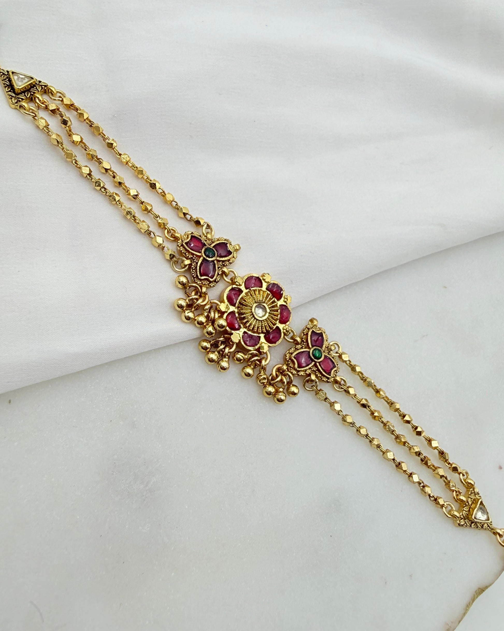 Gold-toned traditional Indian jewelry piece featuring a central floral motif embellished with red and purple stones, resting on a draped white fabric background.