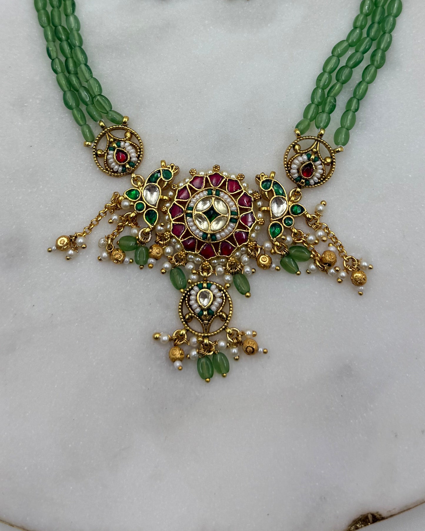 Gold-plated Indian traditional jewelry set featuring a necklace and matching earrings with green beads, red stones, and white pearls, displayed on a white marble surface.
