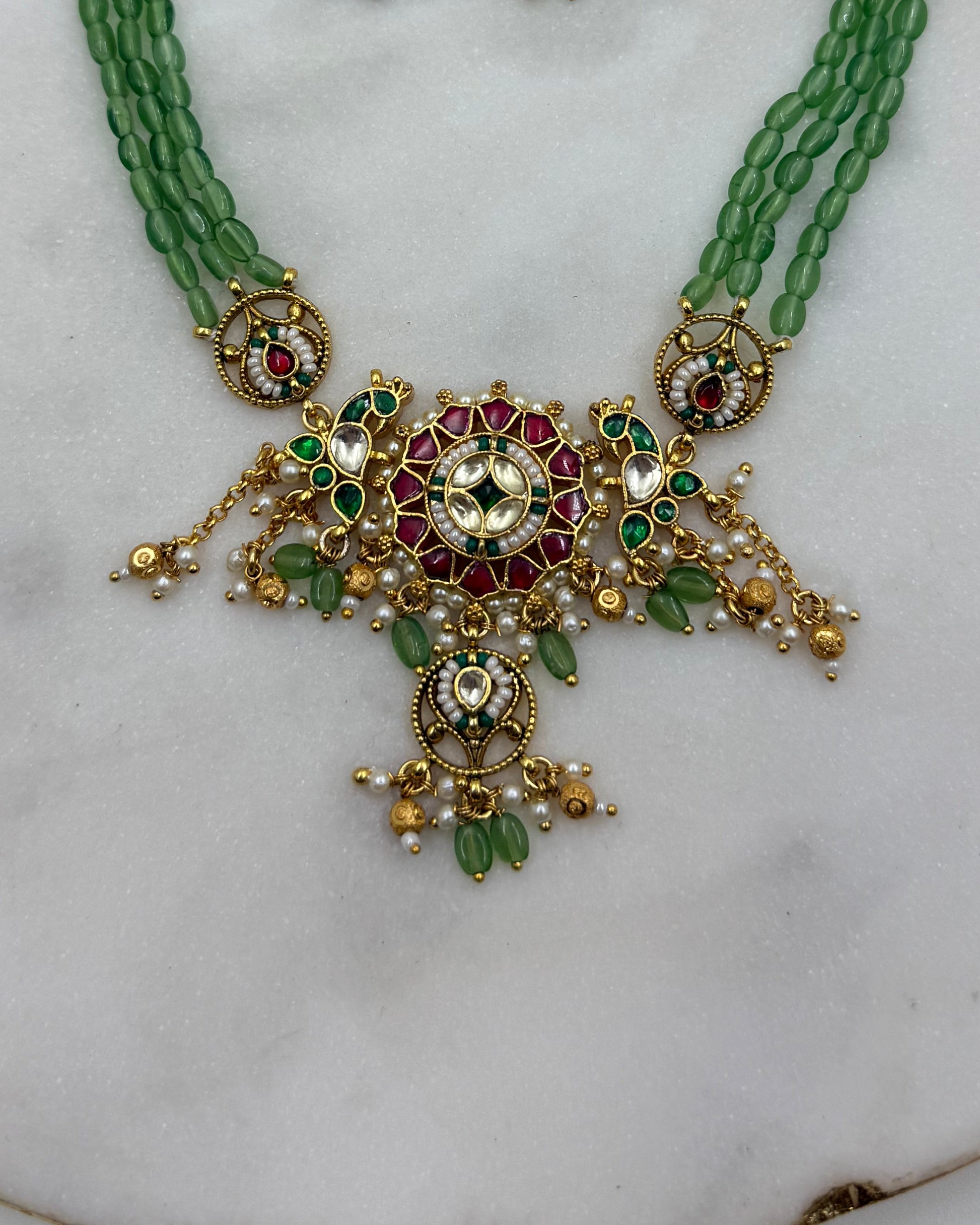Gold-plated Indian traditional jewelry set featuring a necklace and matching earrings with green beads, red stones, and white pearls, displayed on a white marble surface.