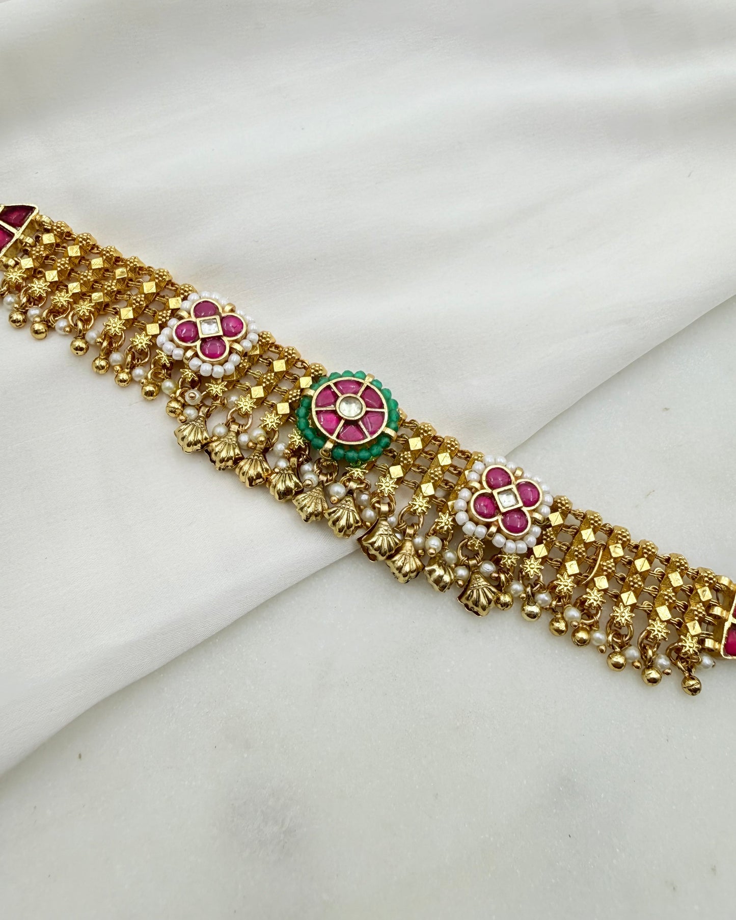 Gold-toned traditional Indian jewelry belt with floral enameled motifs in pink, green, and white, displayed on a draped white fabric background.