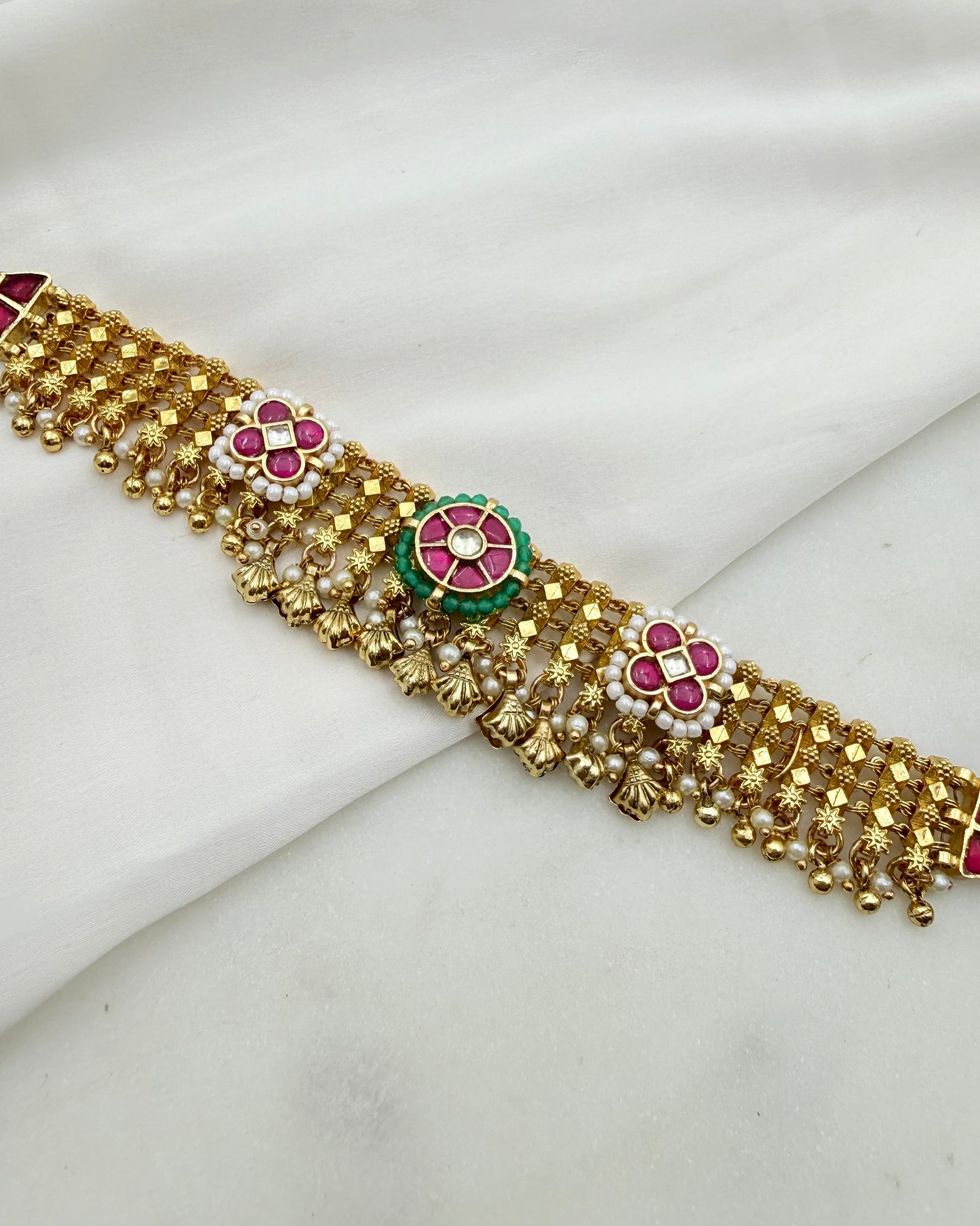 Gold-toned traditional Indian jewelry belt with floral enameled motifs in pink, green, and white, displayed on a draped white fabric background.