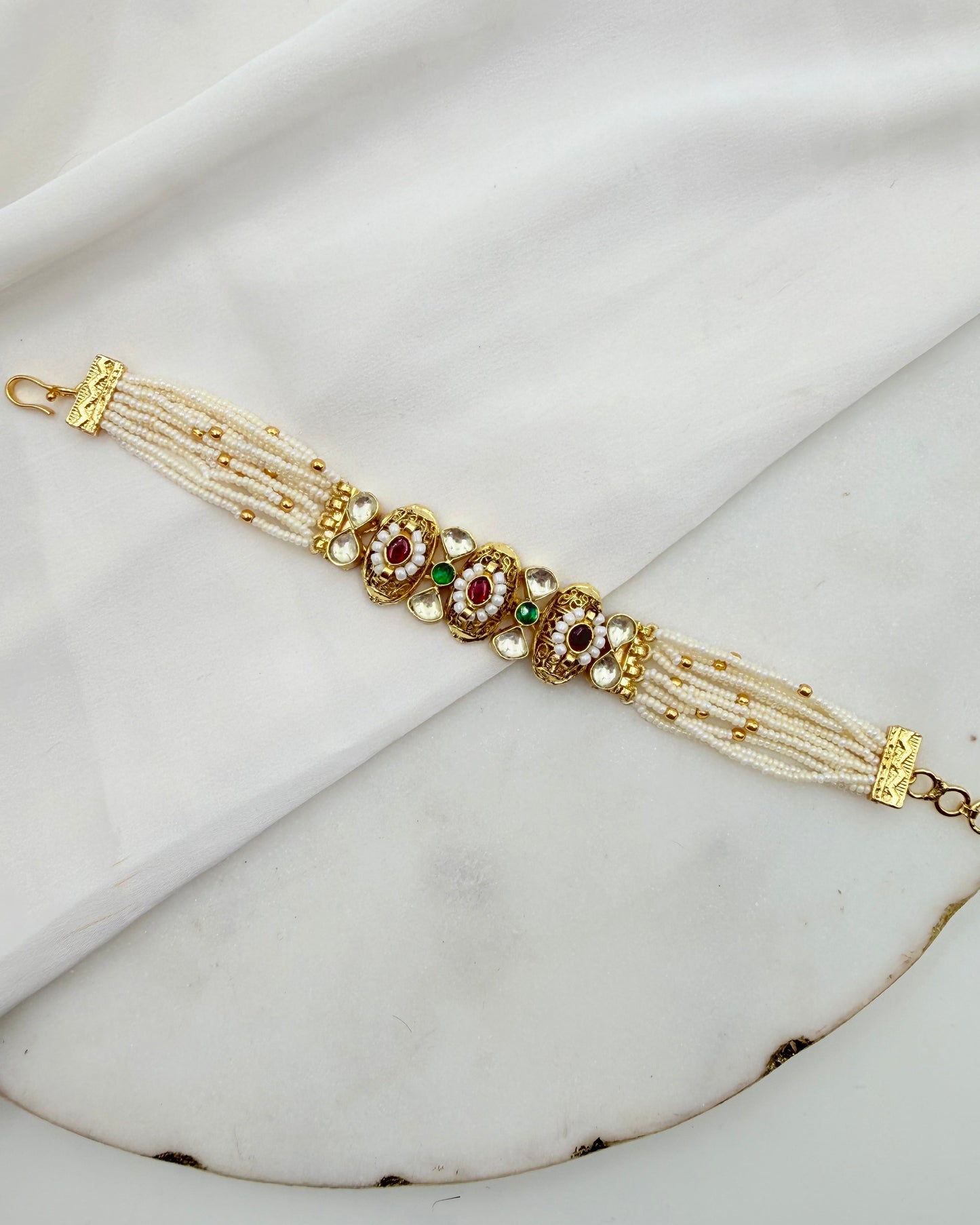 A traditional Indian Kundan bracelet with white pearl-like strands, gold-plated metal, and red and green stone embellishments, displayed on a white marble surface with a white fabric backdrop.