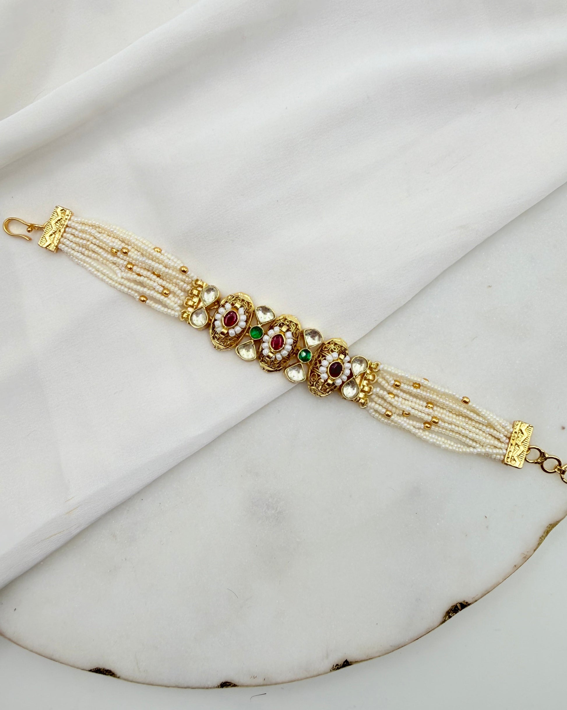 A traditional Indian Kundan bracelet with white pearl-like strands, gold-plated metal, and red and green stone embellishments, displayed on a white marble surface with a white fabric backdrop.