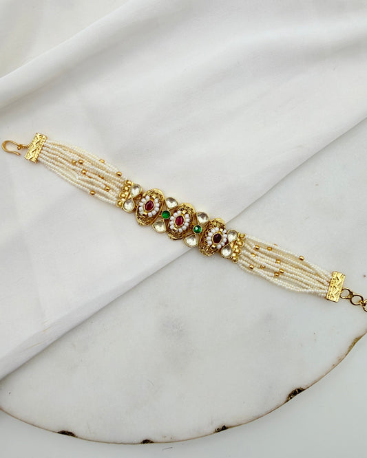 A traditional Indian Kundan bracelet with white pearl-like strands, gold-plated metal, and red and green stone embellishments, displayed on a white marble surface with a white fabric backdrop.