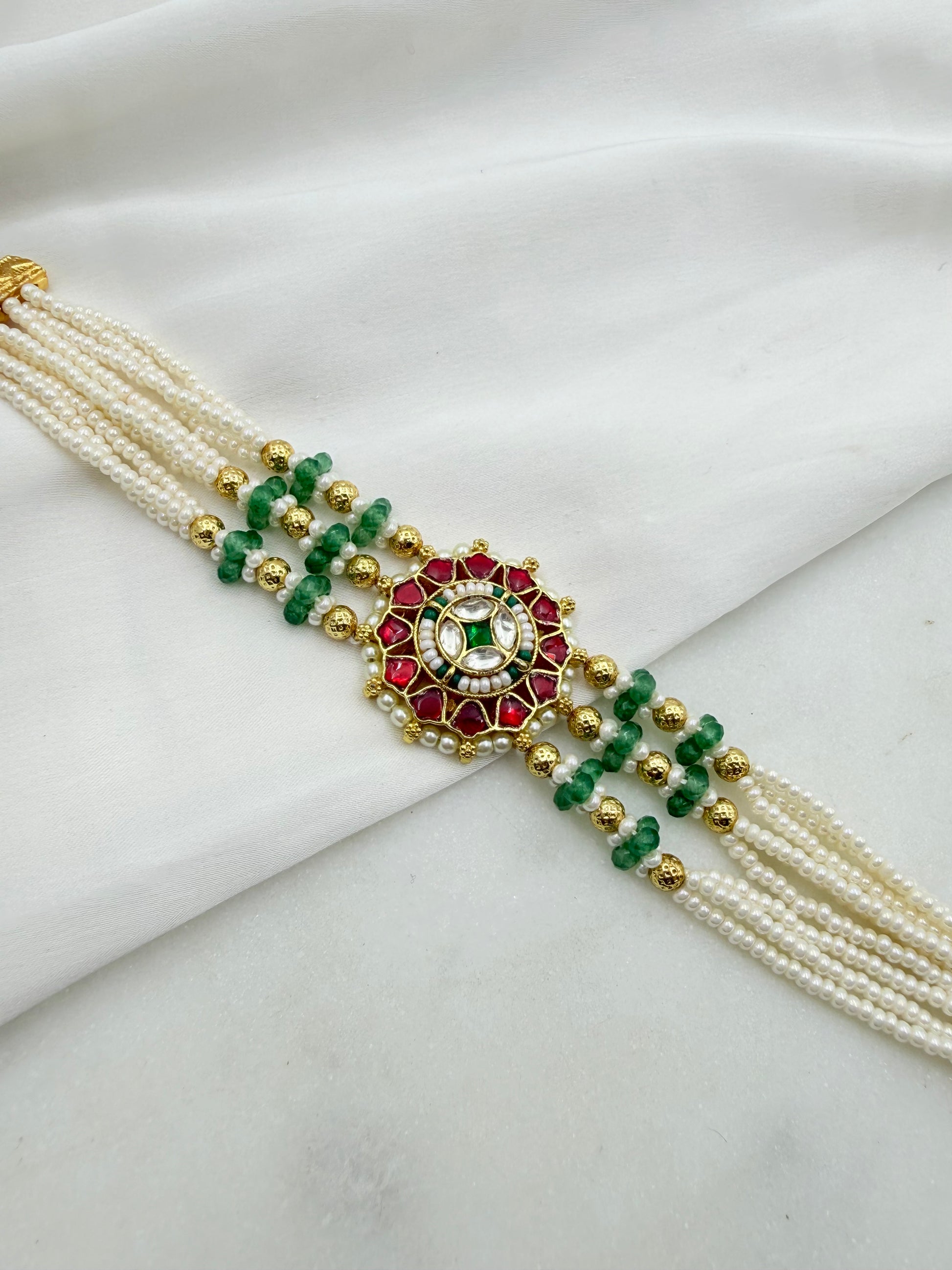 A traditional Indian multi-strand pearl bracelet (silk bracelet) featuring a central gold-plated mandarin ornament with red and green enamel, set against a draped white fabric background.