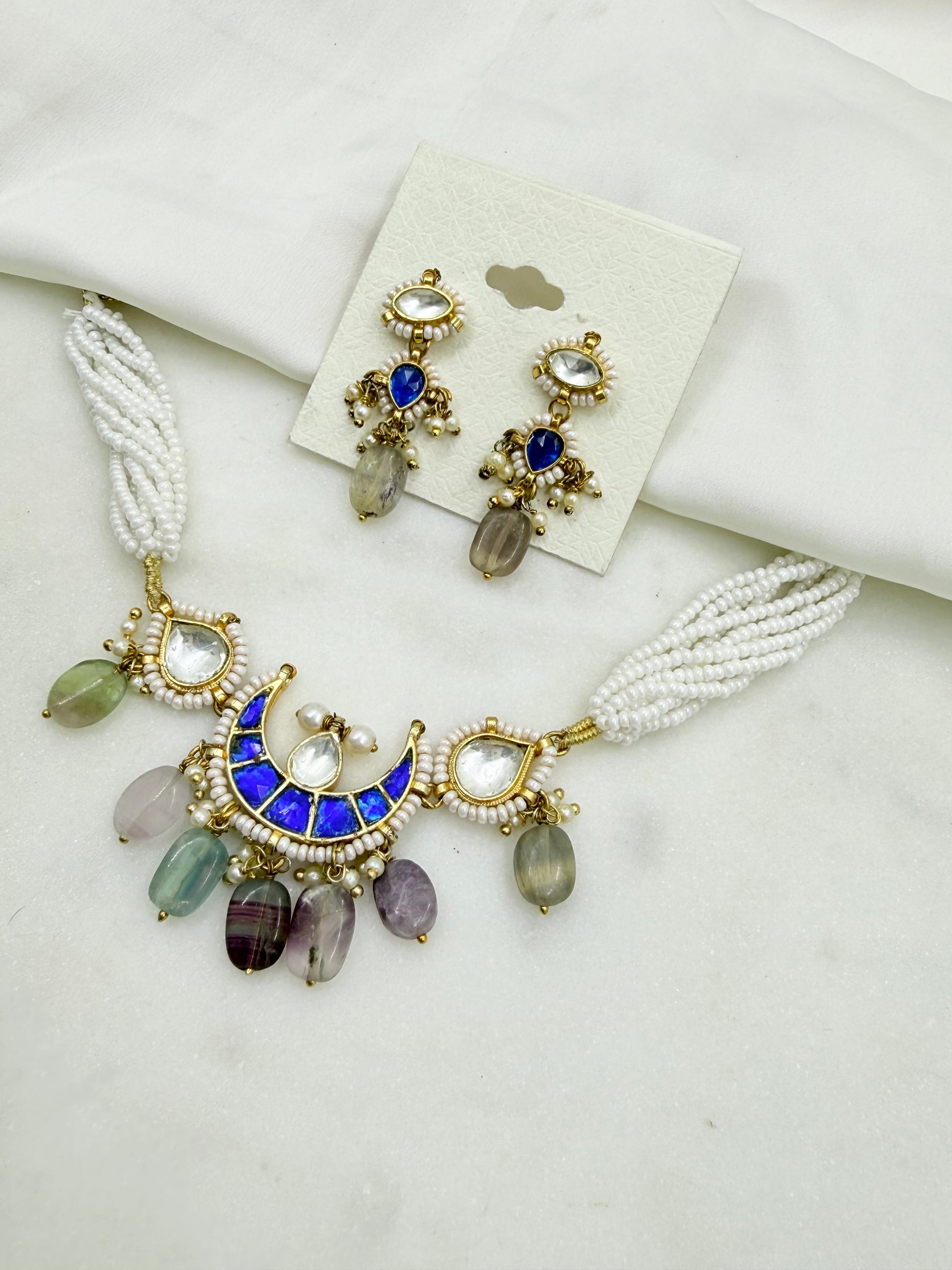 A gold-plated jewelry set featuring a statement necklace and matching drop earrings. The necklace has white beaded strands, a gold crescent moon centerpiece with a blue enamel inlay, and dangling faceted beads in green, purple, and gray.