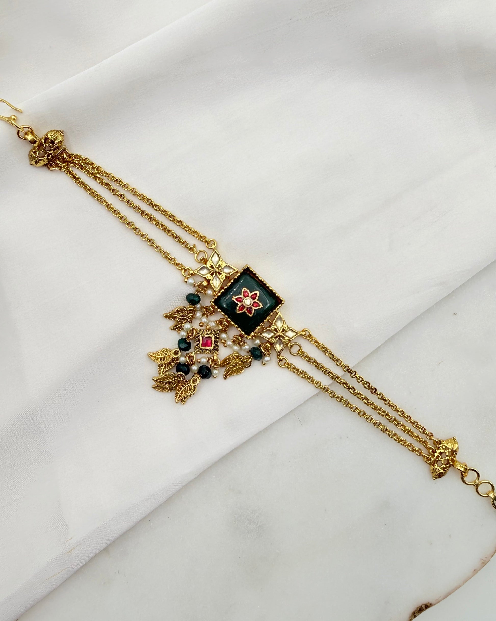 A gold-plated traditional Indian choker necklace with a central square black enamel panel featuring a red and white floral motif, adorned with hanging gold leaf charms, green beads, and small pearl-like accents, displayed on a white marble surface.