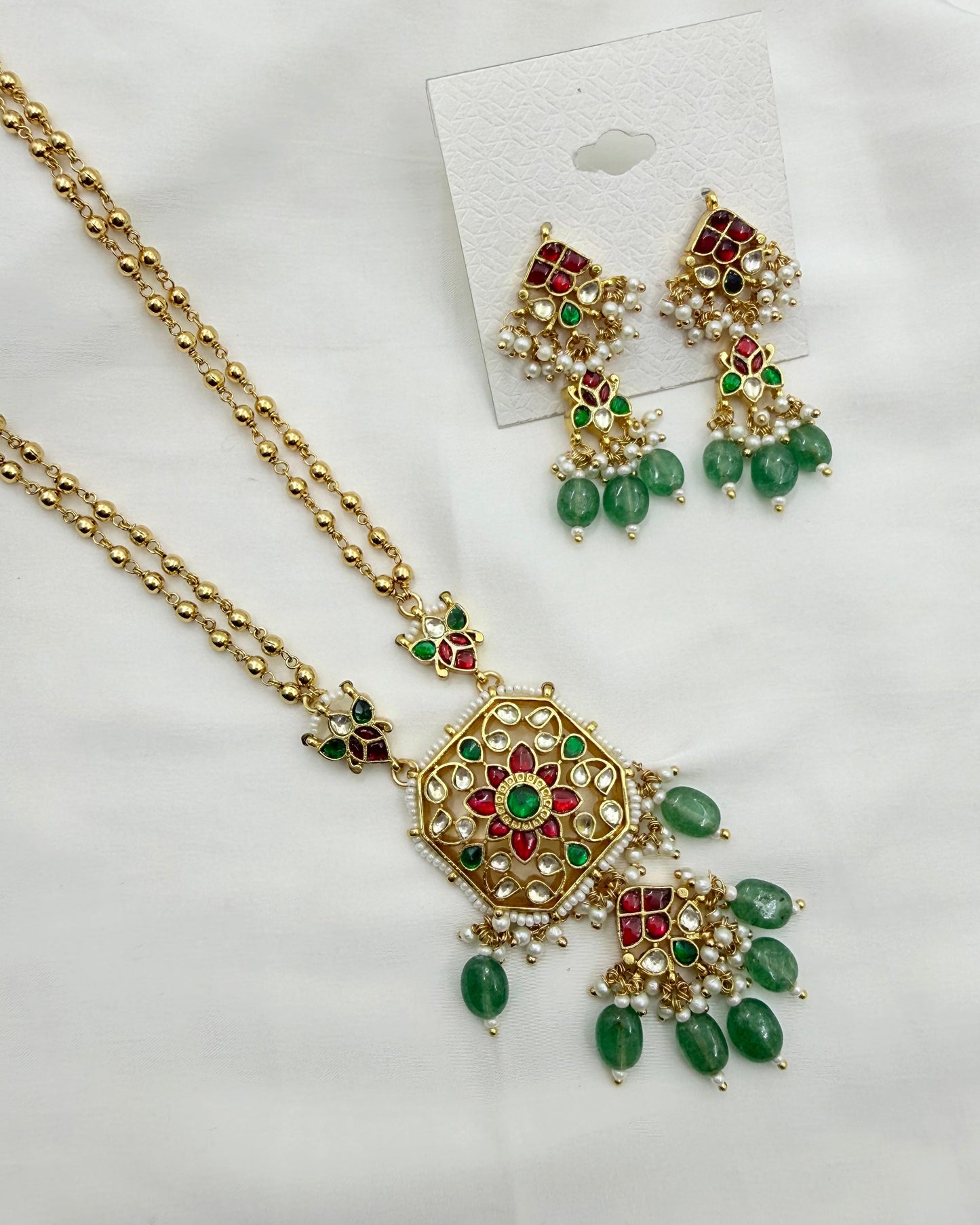 Gold-plated Indian traditional jewelry set featuring a necklace and matching earrings with green stones, red stones, and white faux pearls, displayed on a white fabric background.