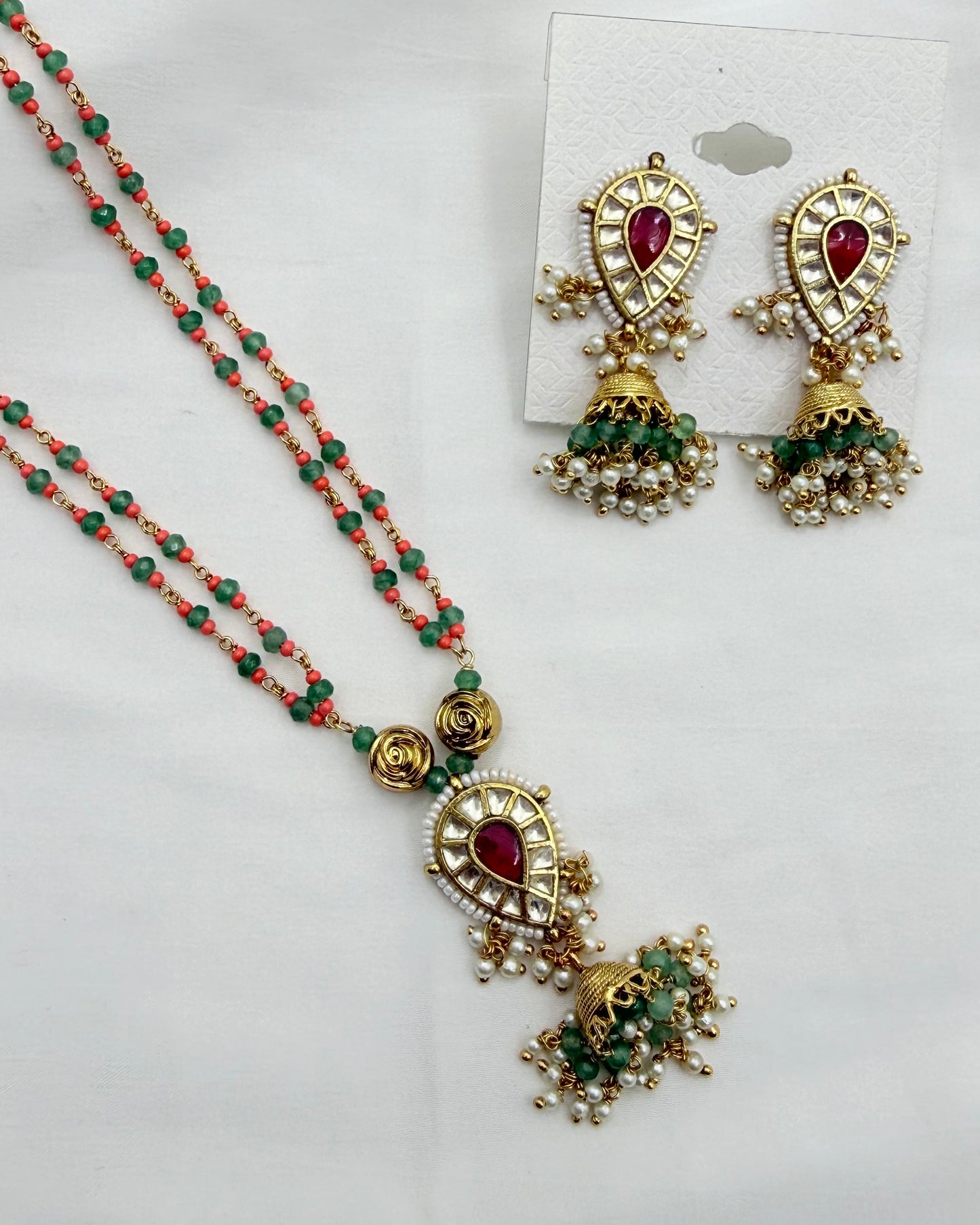 Gold-plated traditional Indian jewelry set featuring a beaded necklace and matching earrings with red stones and green bead accents, displayed on a white fabric background.