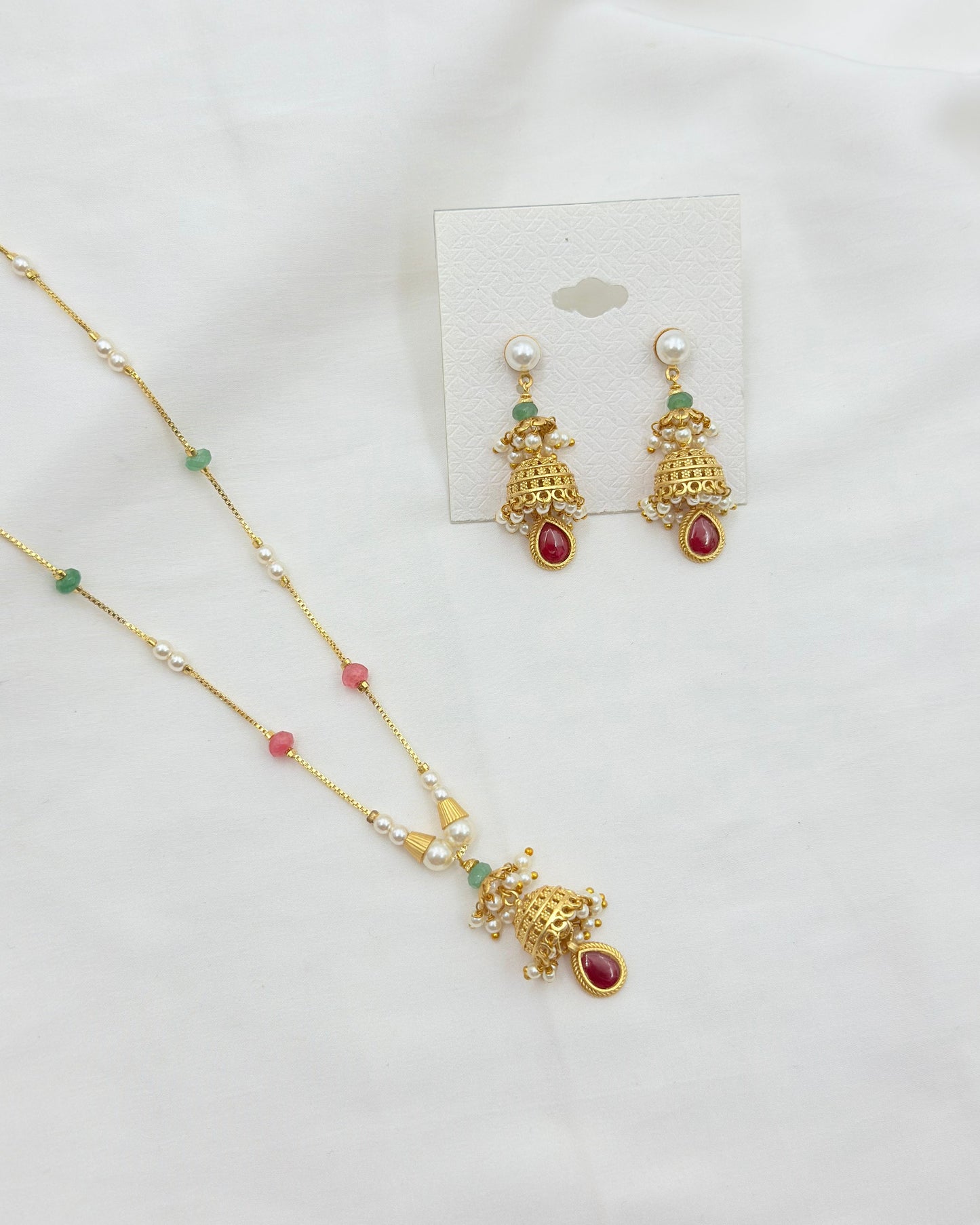 Gold-toned traditional Indian jewelry set featuring a necklace and matching earrings with white pearls, green stones, and red gemstone accents, displayed on a white fabric background.
