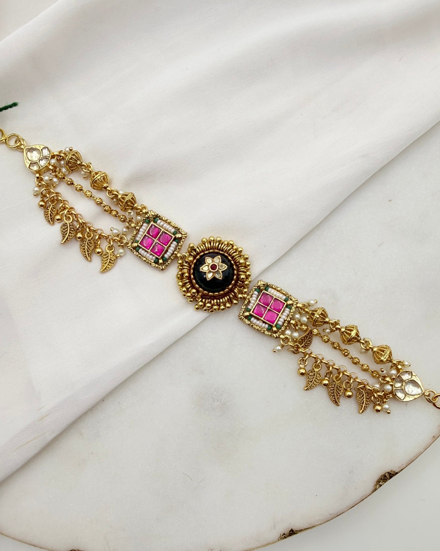 Gold-toned traditional Indian jewelry piece featuring a central black oval medallion with a floral motif, flanked by two square pink and green enamel panels and dangling gold leaf and chain elements, displayed on a white fabric background.
