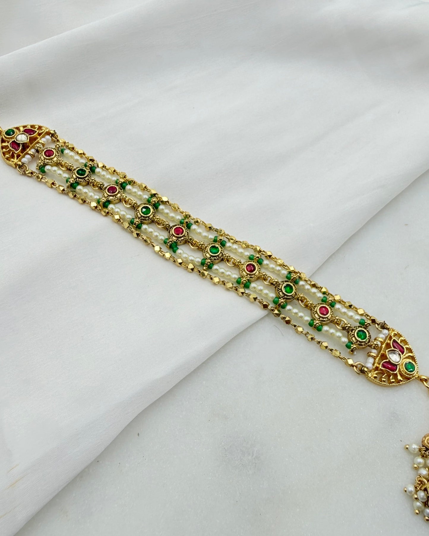 An ornate gold-plated traditional Indian bracelet (choker) decorated with white pearls, green and red enamel stones, and a dangling gold charm, displayed on a draped white fabric background.