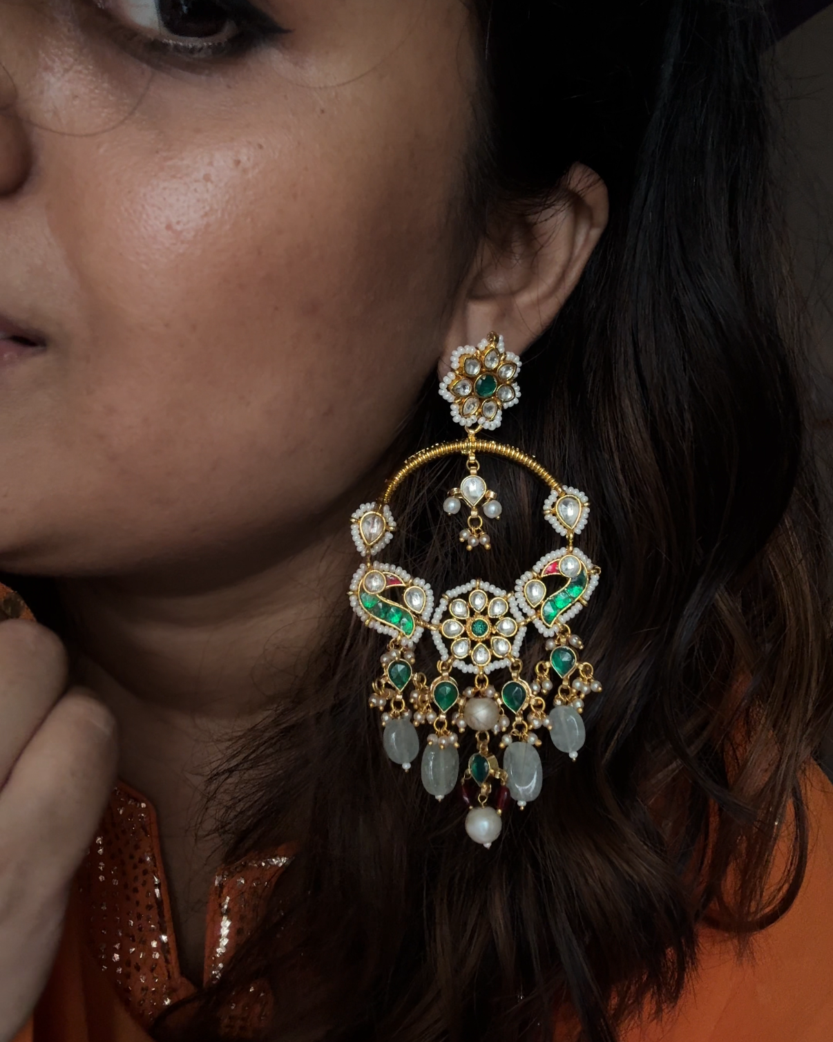 A close-up lifestyle shot of a woman wearing a large, ornate gold-toned dangle earring decorated with green and white stones.