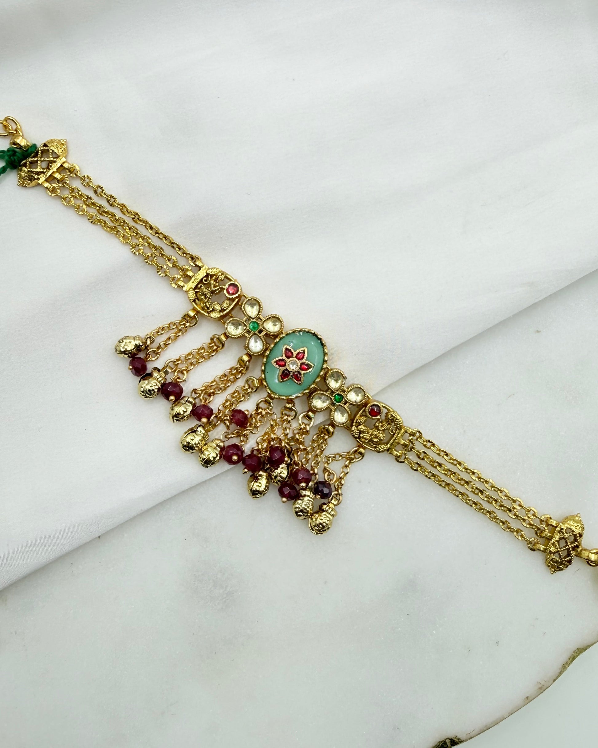 Gold-toned traditional Indian jewelry piece featuring a central oval turquoise enamel medallion with a red floral motif, surrounded by small red and green stones, and multiple dangling chain elements with gold beads.