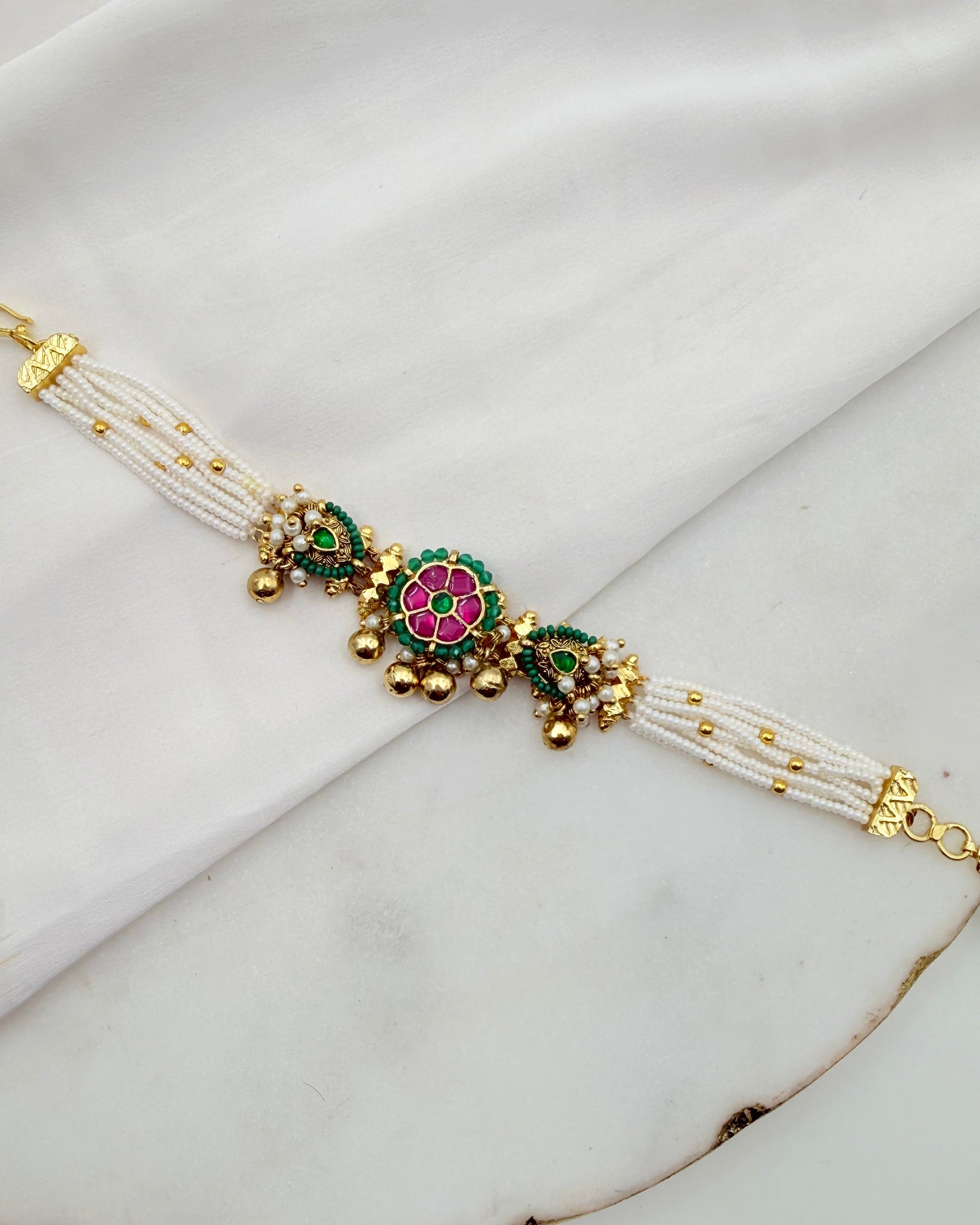 A traditional Indian multi-strand pearl bracelet with gold-plated metal details, featuring a central pink and green floral motif with small gold bells, displayed on a white fabric background.