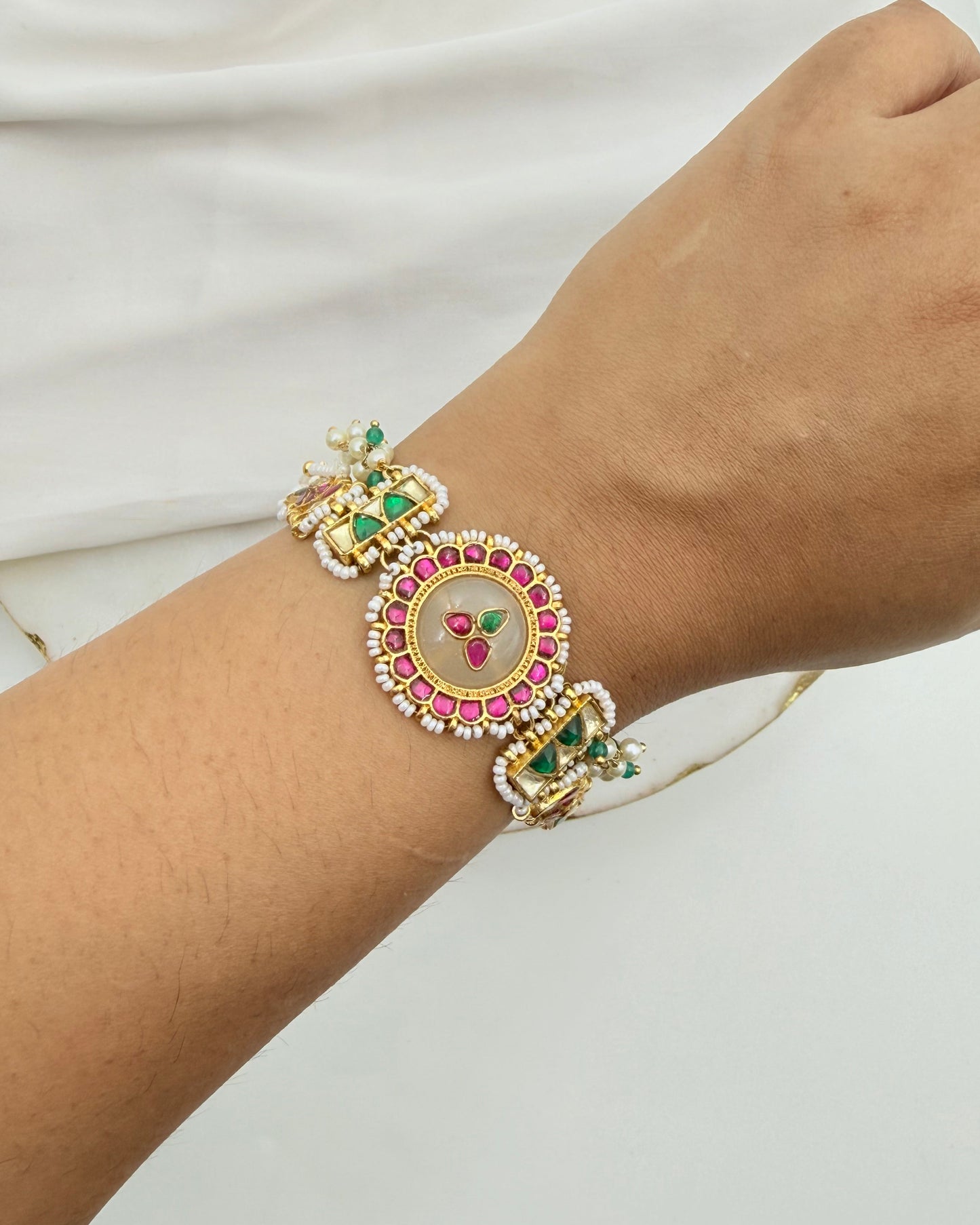 A close-up of a person's wrist wearing a highly detailed, ornate gold-plated traditional Indian bracelet (Kara) decorated with pink and green stones and white crystal-like accents.