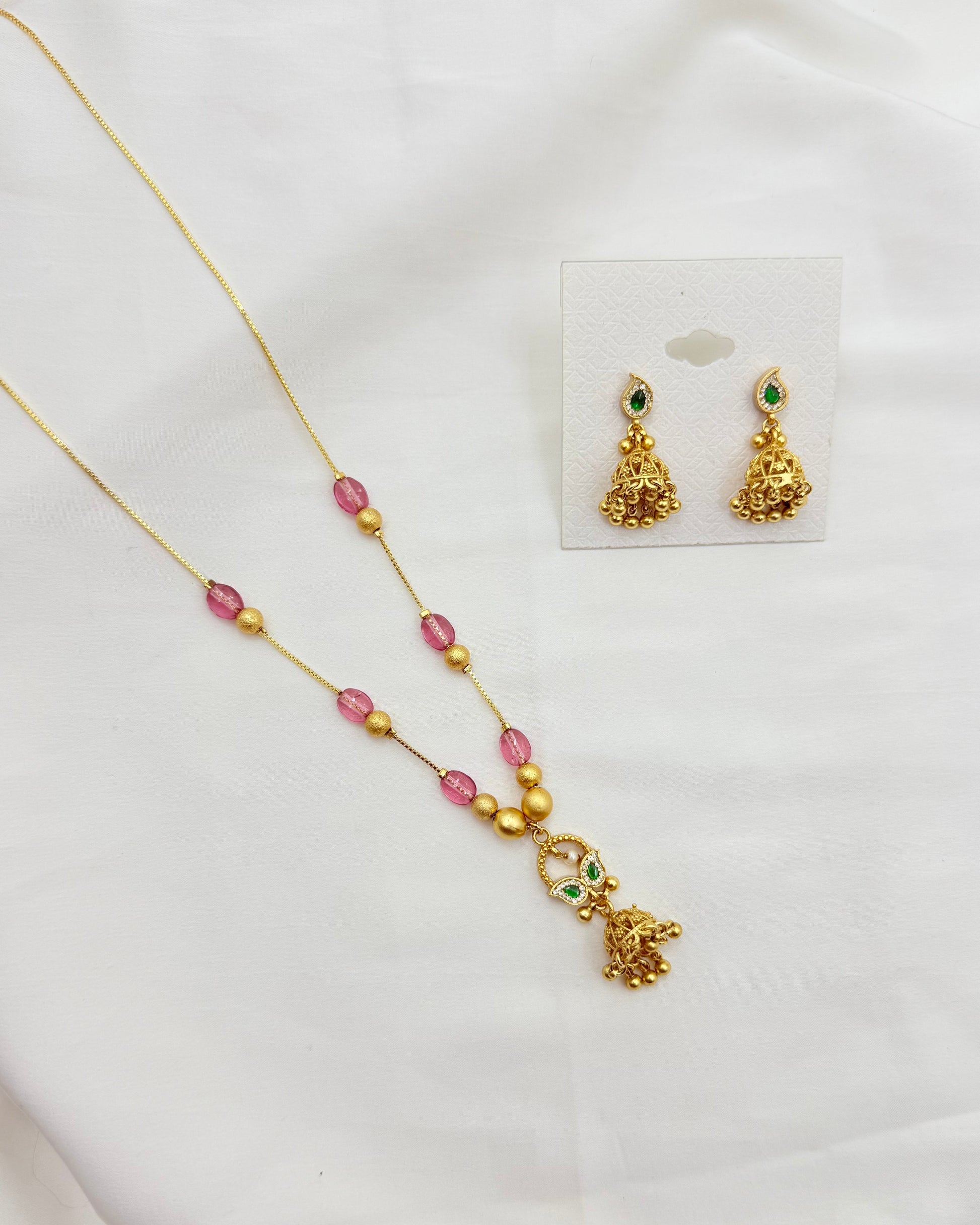 Gold-toned jewelry set on a white fabric background, featuring a necklace and matching earrings. The necklace has pink and gold beads with a decorative gold pendant.