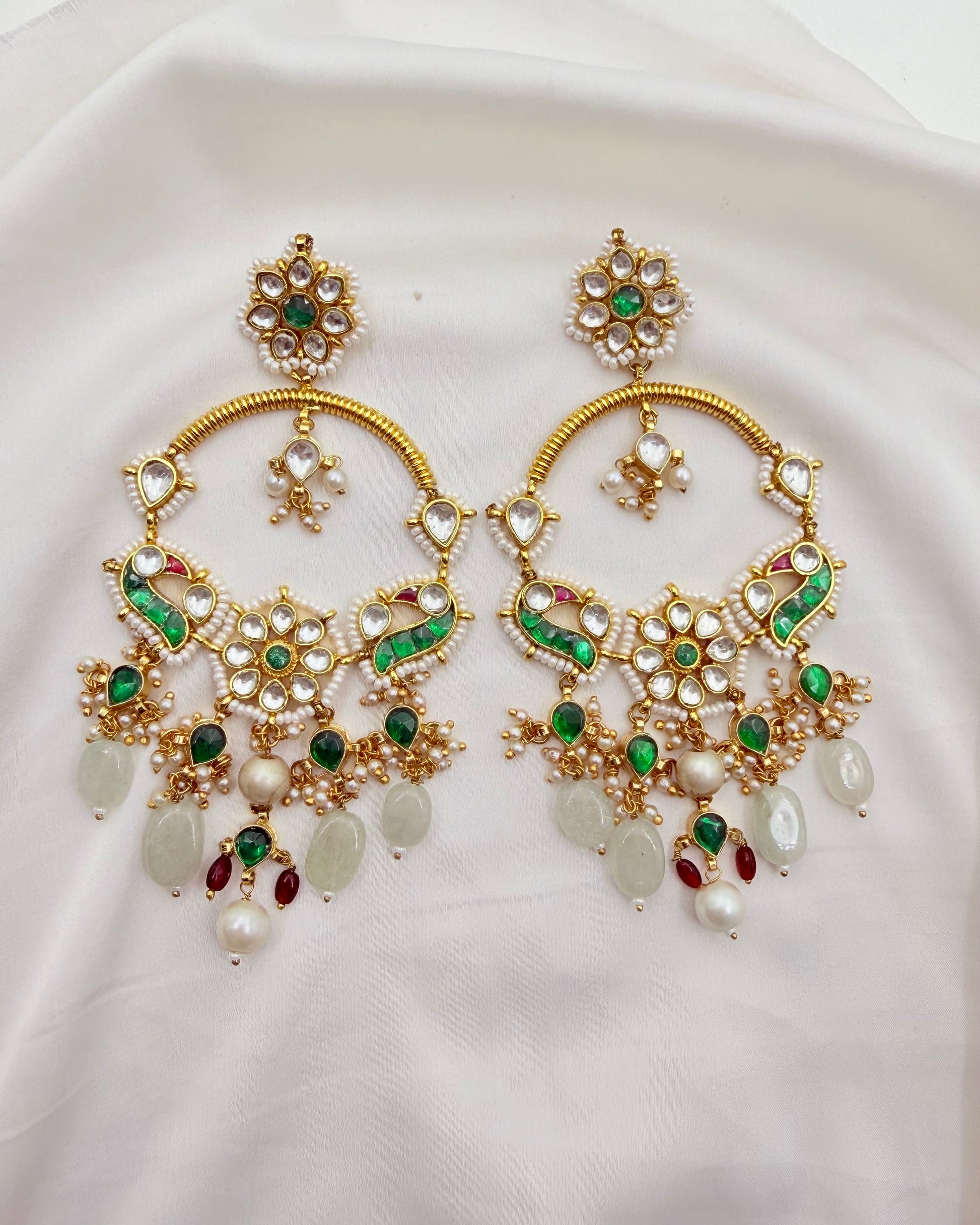 A pair of ornate gold-toned Indian chandelier earrings decorated with white faux pearls, green stones, and red accent stones, displayed on a white fabric background.