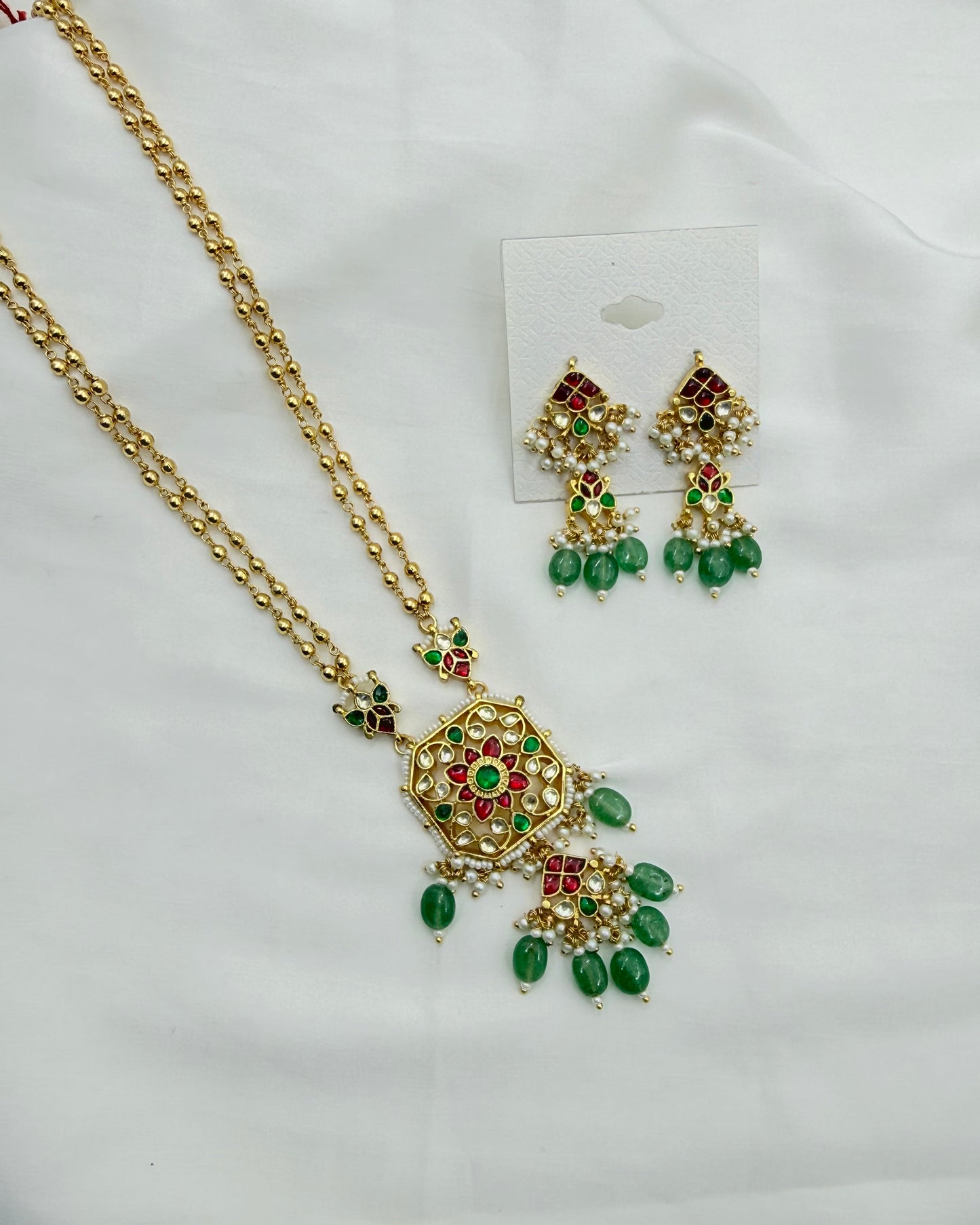 Gold-toned traditional necklace and matching drop earrings with green and red stone embellishments, displayed on a white fabric background with a small white card holding the earrings.
