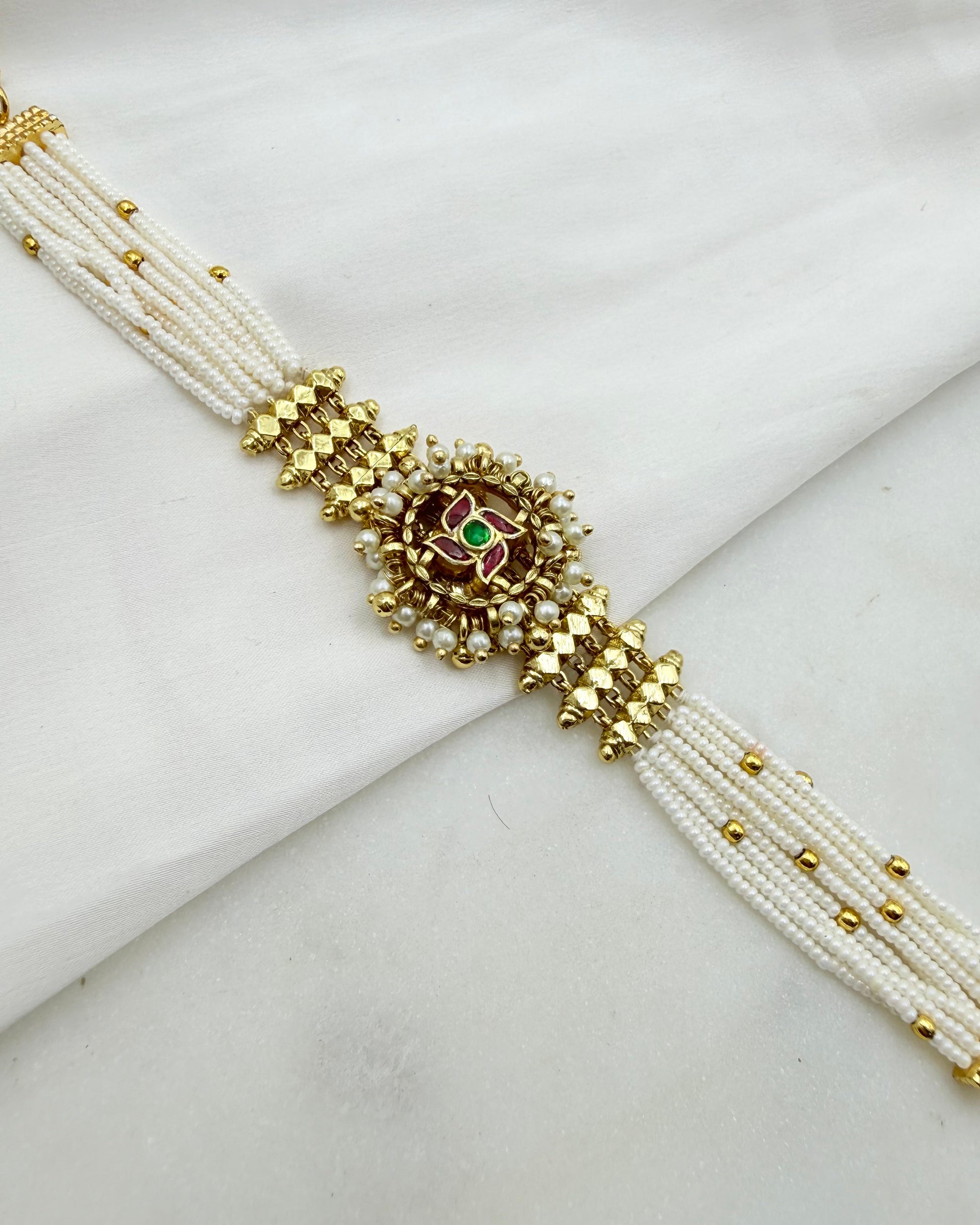 A traditional Indian multi-strand pearl bracelet with gold-plated metal accents, a central floral motif, and a small green stone, displayed on a white fabric background.