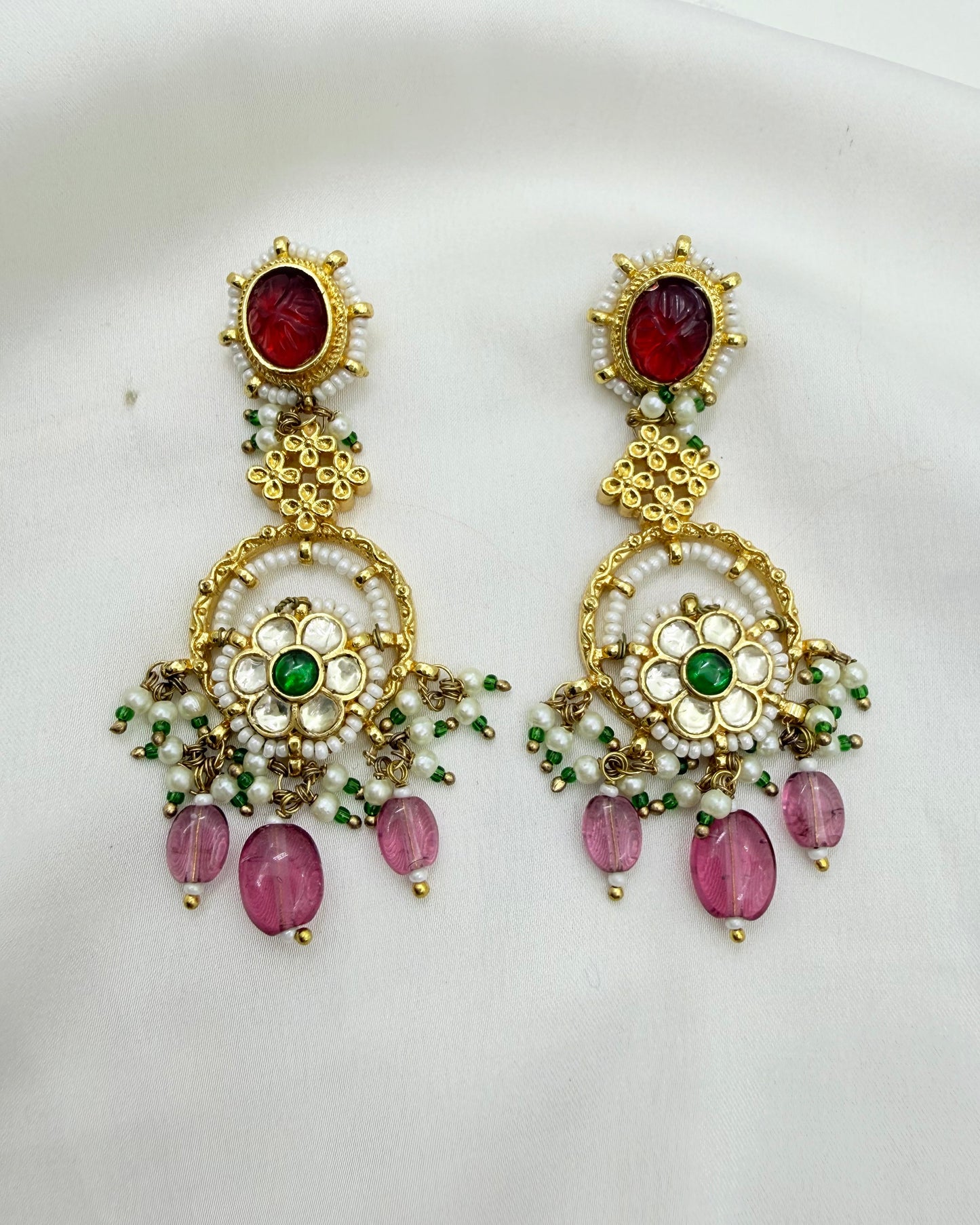 A pair of ornate gold-toned traditional Indian earrings featuring red oval stones, green floral accents, white pearl-like beads, and dangling pink teardrop beads, displayed on a white fabric background.