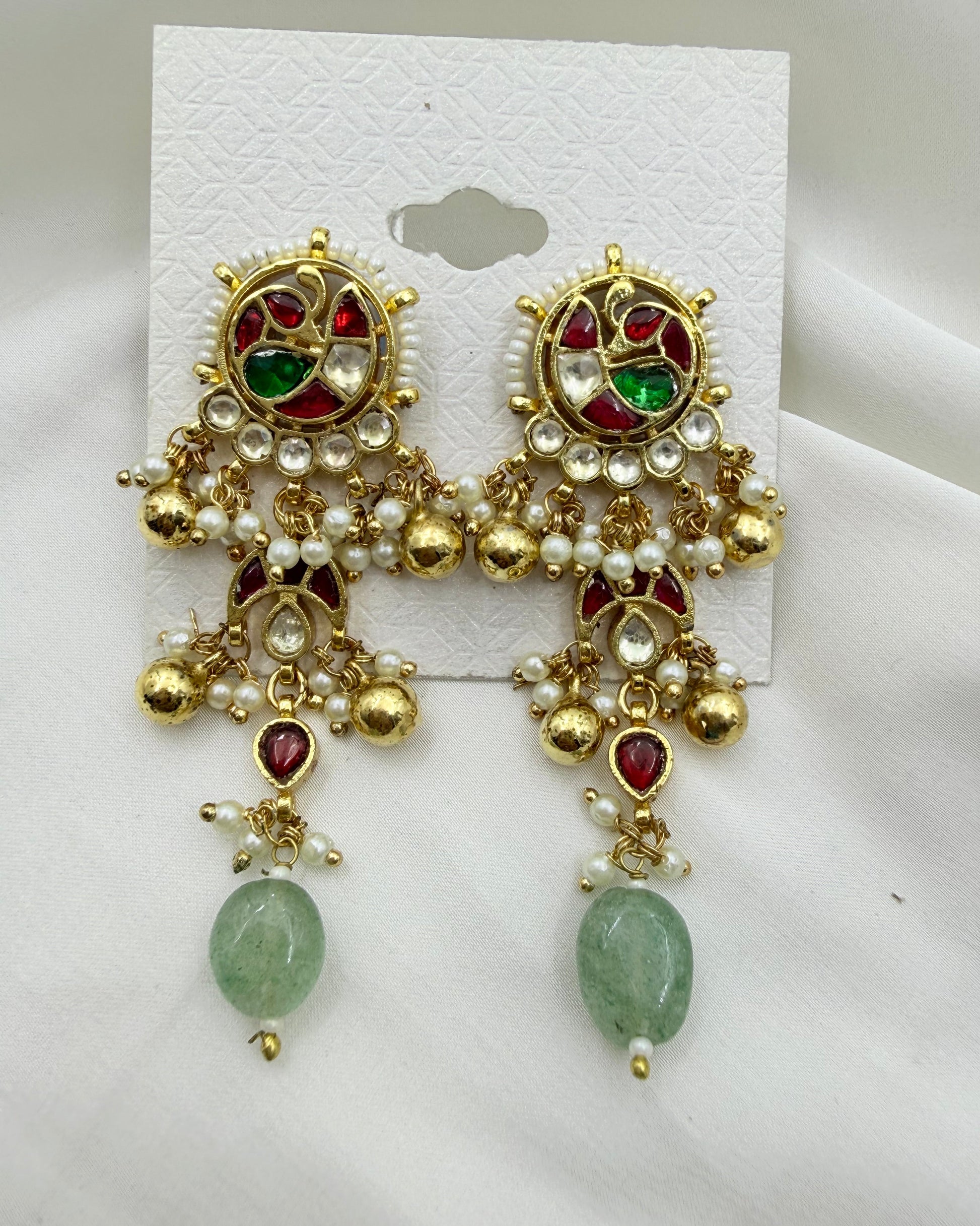 A pair of ornate gold-toned Indian jhumka earrings with red and green stones, white faux pearl beads, and a dangling light green stone, displayed on a white card against a white fabric background.