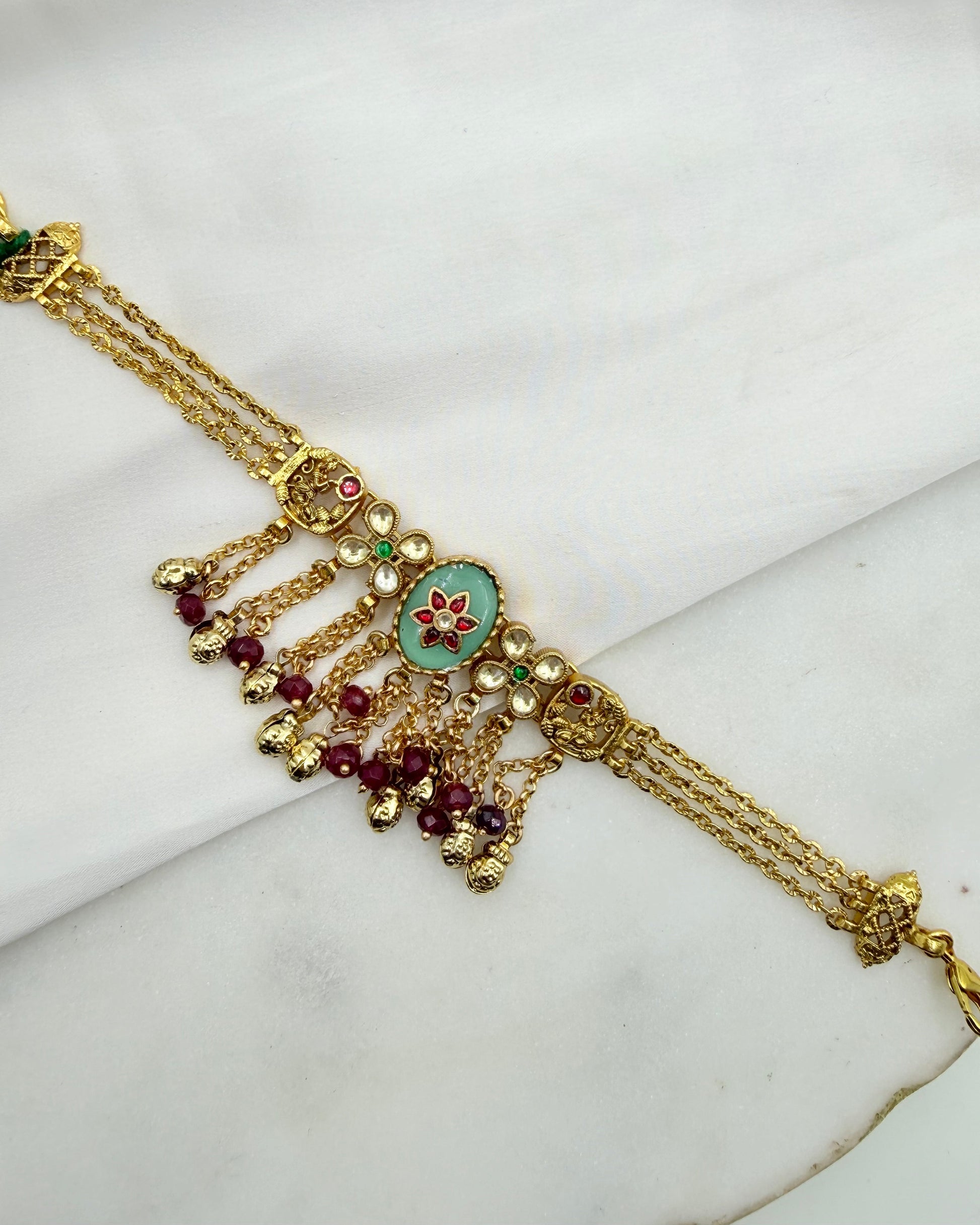 Gold-toned traditional Indian jewelry piece featuring a central oval green enamel motif with red floral detailing, flanked by dangling red bead chains and small gold beads, displayed on a white fabric background.