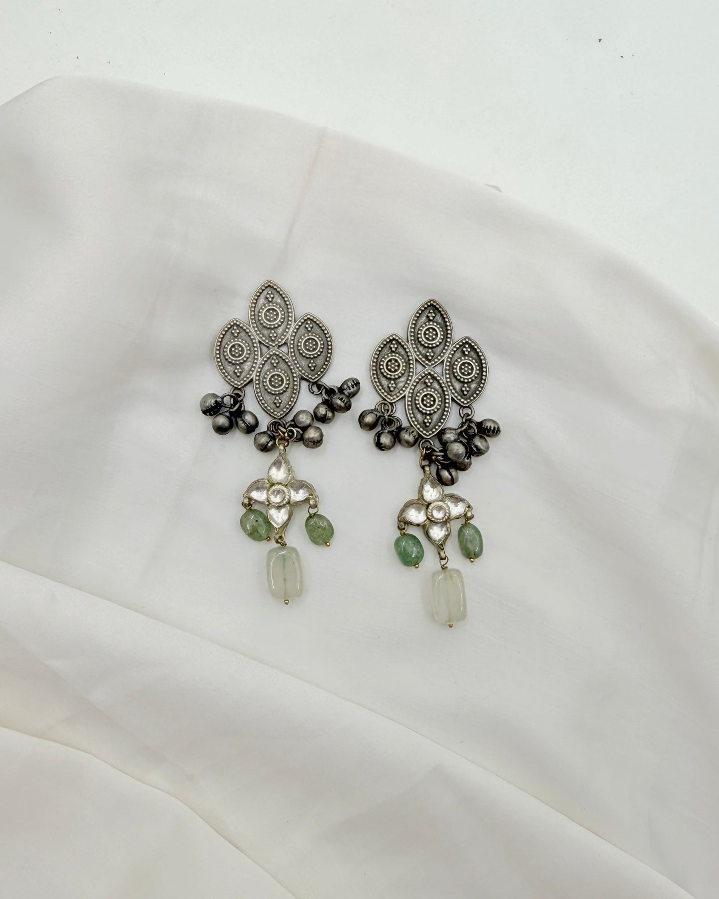 A pair of ornate silver-toned filigree earrings with dark metallic bead accents and green stone drops, displayed on a draped white fabric surface.