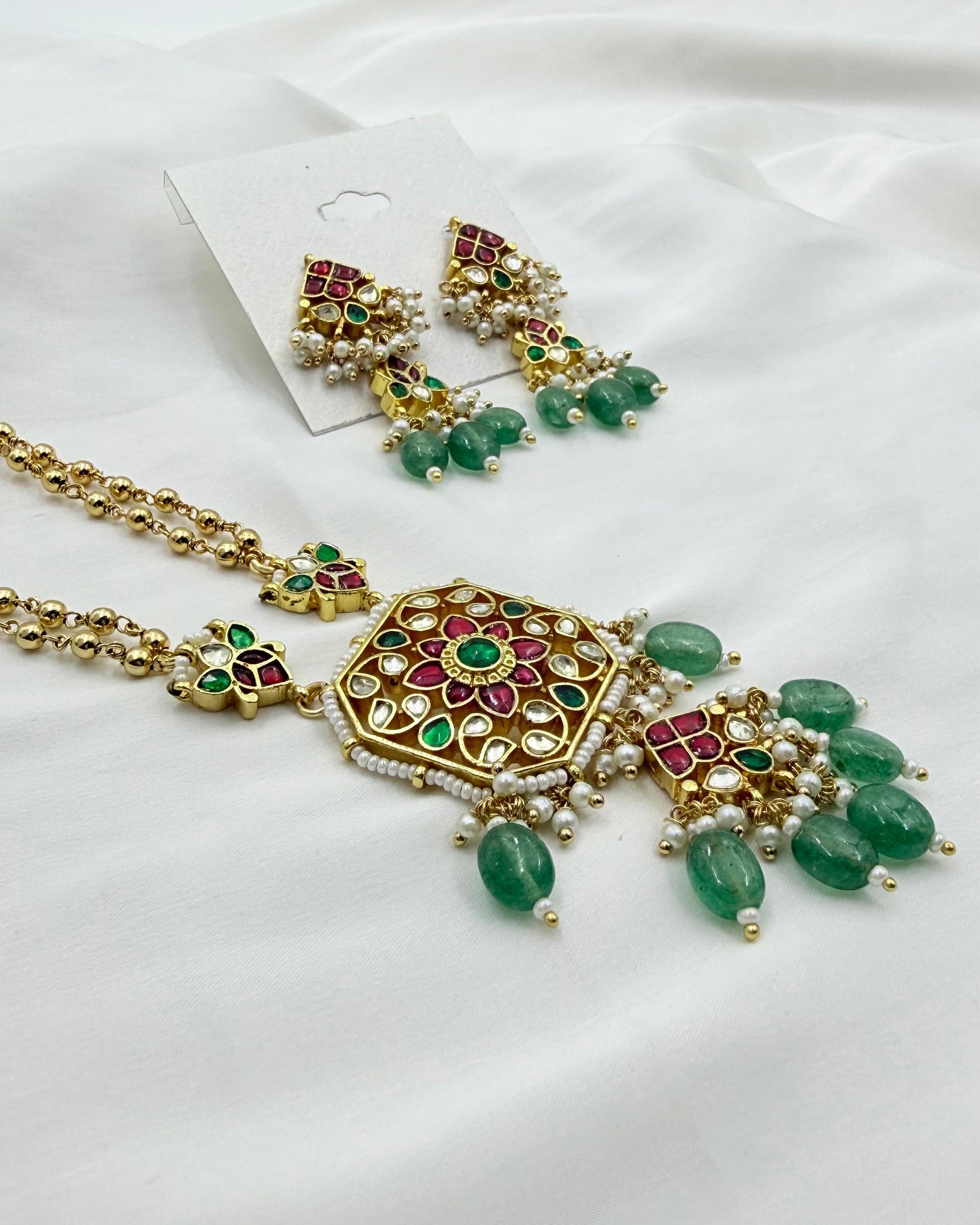 Gold-plated Indian traditional jewelry set featuring a necklace and matching earrings with green stone accents, red enamel details, and white pearl-like beads, displayed on a white fabric background.