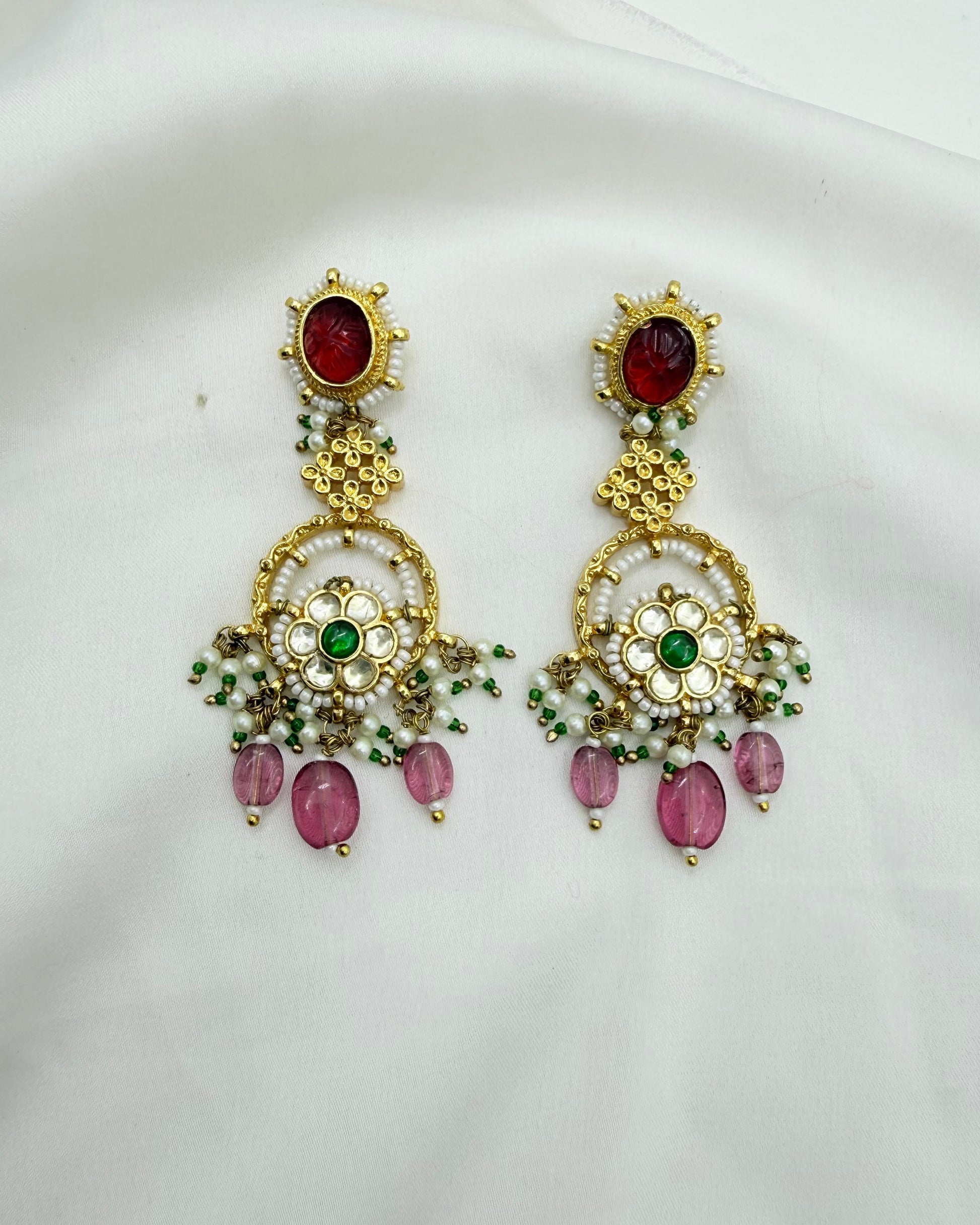 A pair of ornate gold-toned Indian-style earrings with red stones, green accents, and pink dangling beads, displayed on a white fabric background.