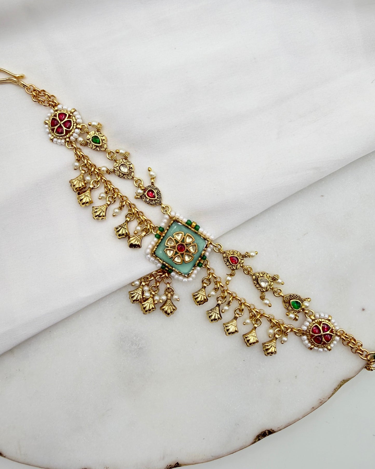An ornate gold-toned traditional Indian jewelry piece, likely a choker or necklace, decorated with a central turquoise-blue square, red and green stone accents, and dangling gold floral elements, displayed on a white marble surface.