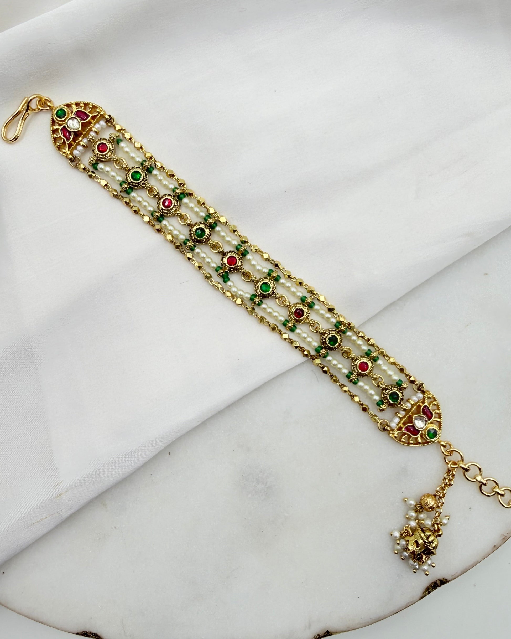 A gold-plated traditional Indian ankle bracelet (bangle) decorated with white faux pearls, green and red enamel details, and a dangling gold charm, displayed on a white fabric background.
