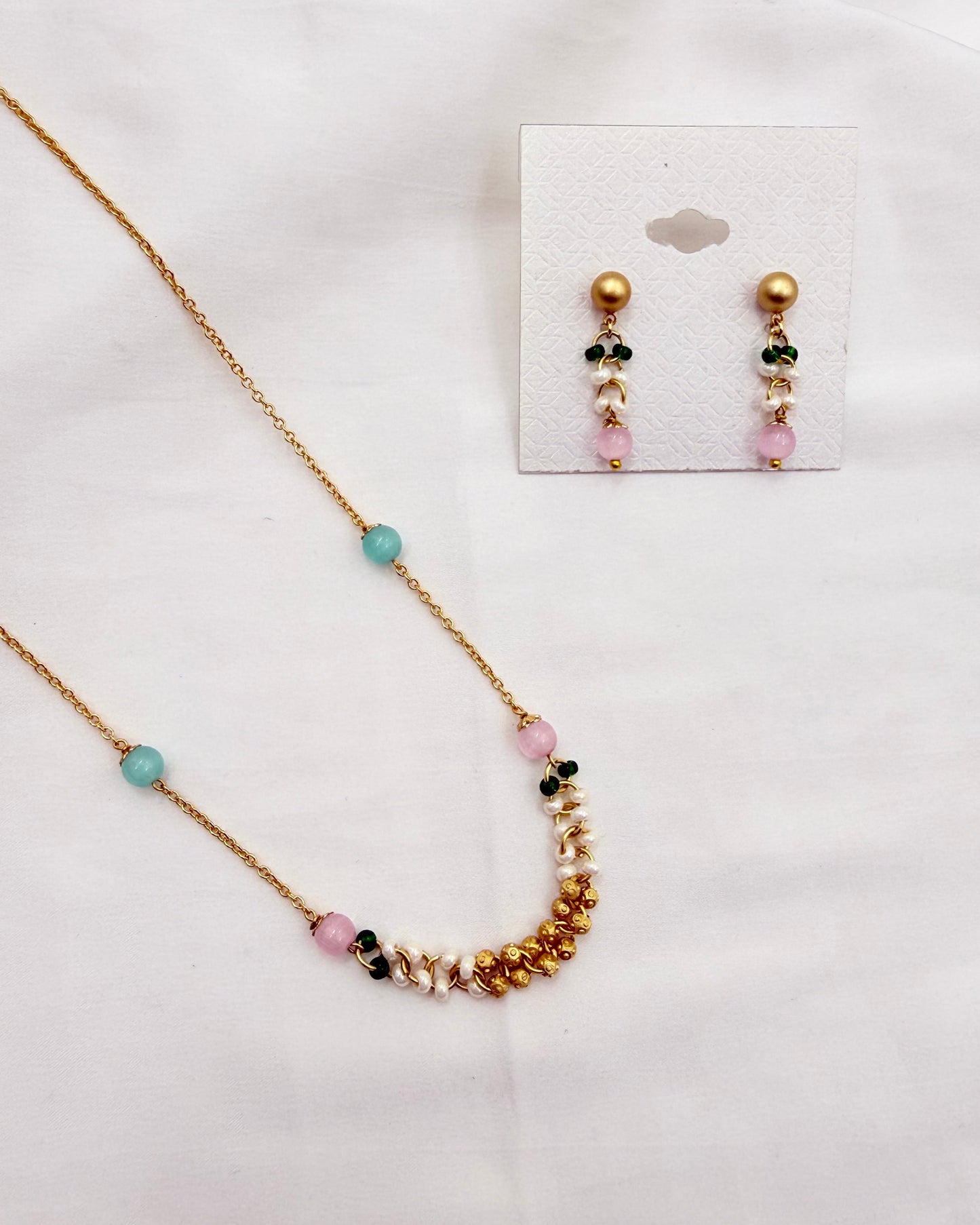 A gold-tone necklace and matching drop earrings displayed on a white fabric background. The necklace has a delicate chain with scattered light blue beads, pink beads, and a curved section featuring gold beads, white pearl-like beads, and small dark green and pink beads.