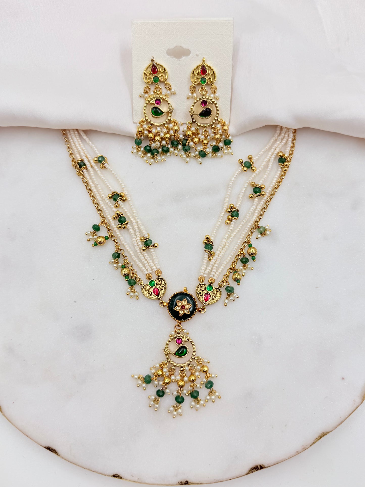 An ornate Indian-style jewelry set featuring a multi-strand necklace and matching earrings with gold-plated details, white faux pearls, green stones, and red accents, displayed on a white marble surface.