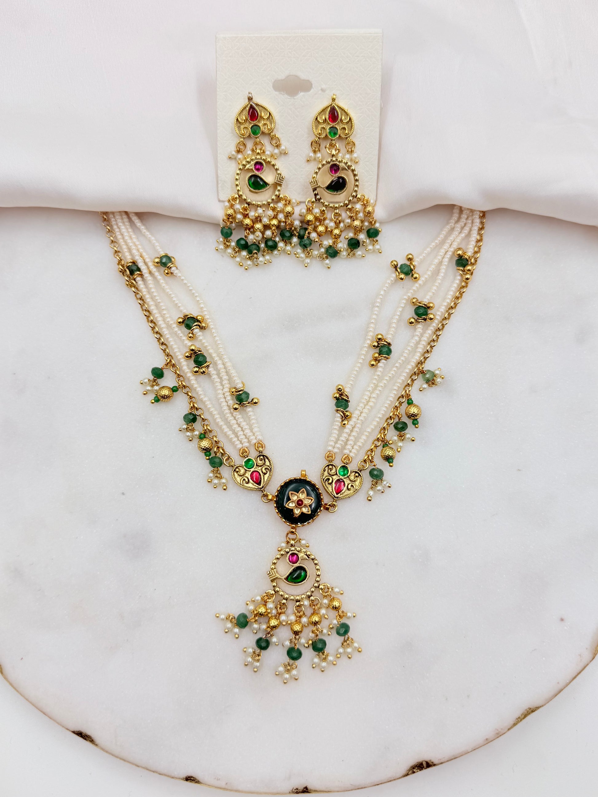 An ornate Indian-style jewelry set featuring a multi-strand necklace and matching earrings with gold-plated details, white faux pearls, green stones, and red accents, displayed on a white marble surface.