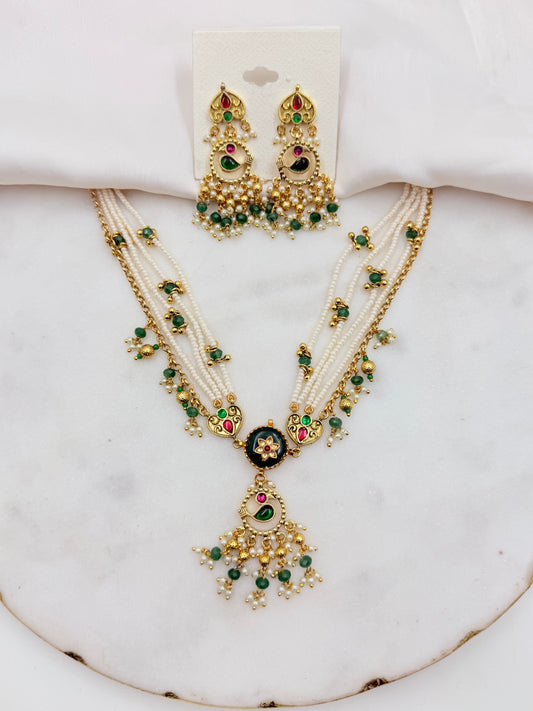 An ornate Indian-style jewelry set featuring a multi-strand necklace and matching earrings with gold-plated details, white faux pearls, green stones, and red accents, displayed on a white marble surface.