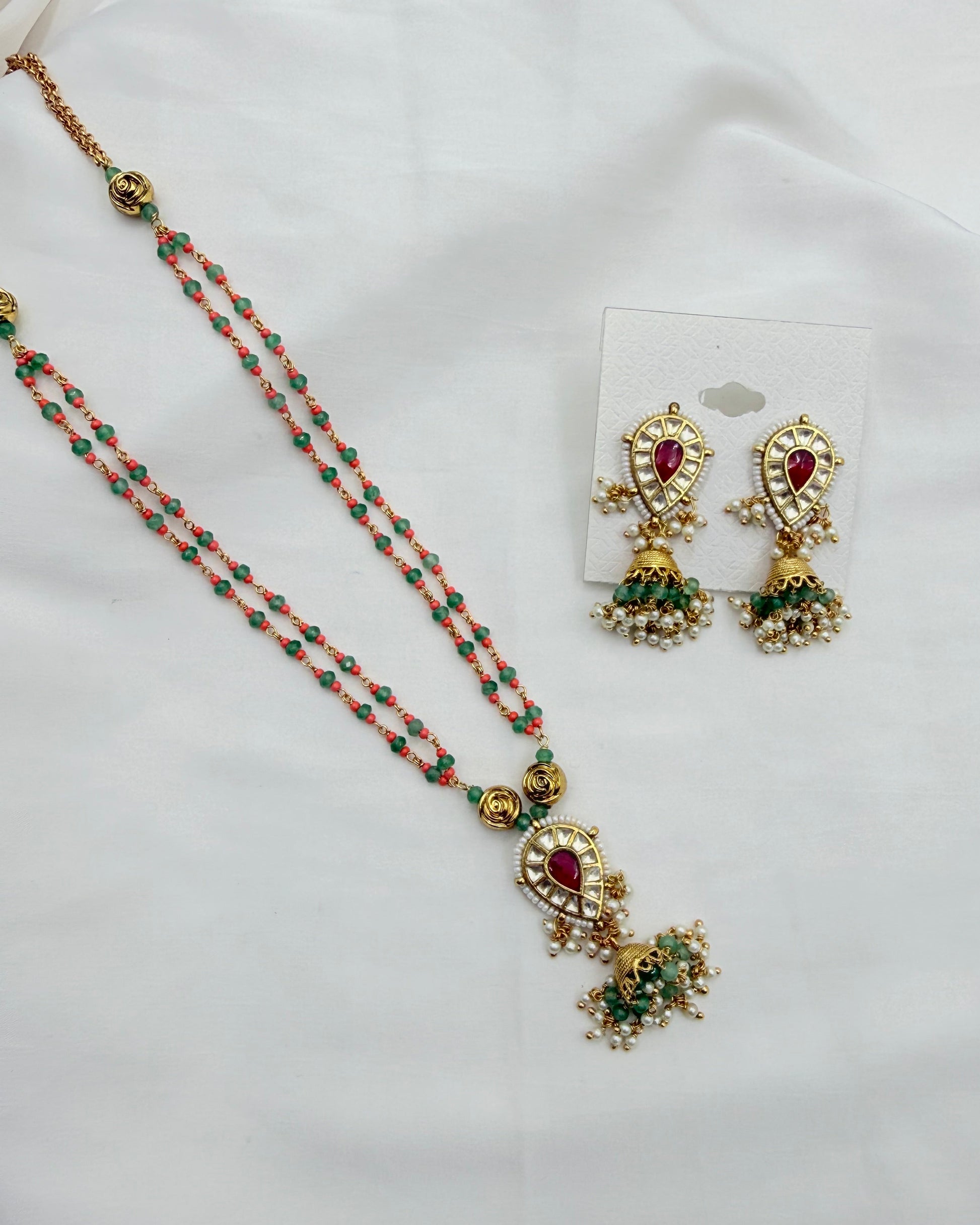 A traditional Indian jewelry set featuring a double-strand necklace and matching earrings. The necklace has coral-pink and green beads with gold-plated elements and a red teardrop stone, while the matching earrings are on a small white card.