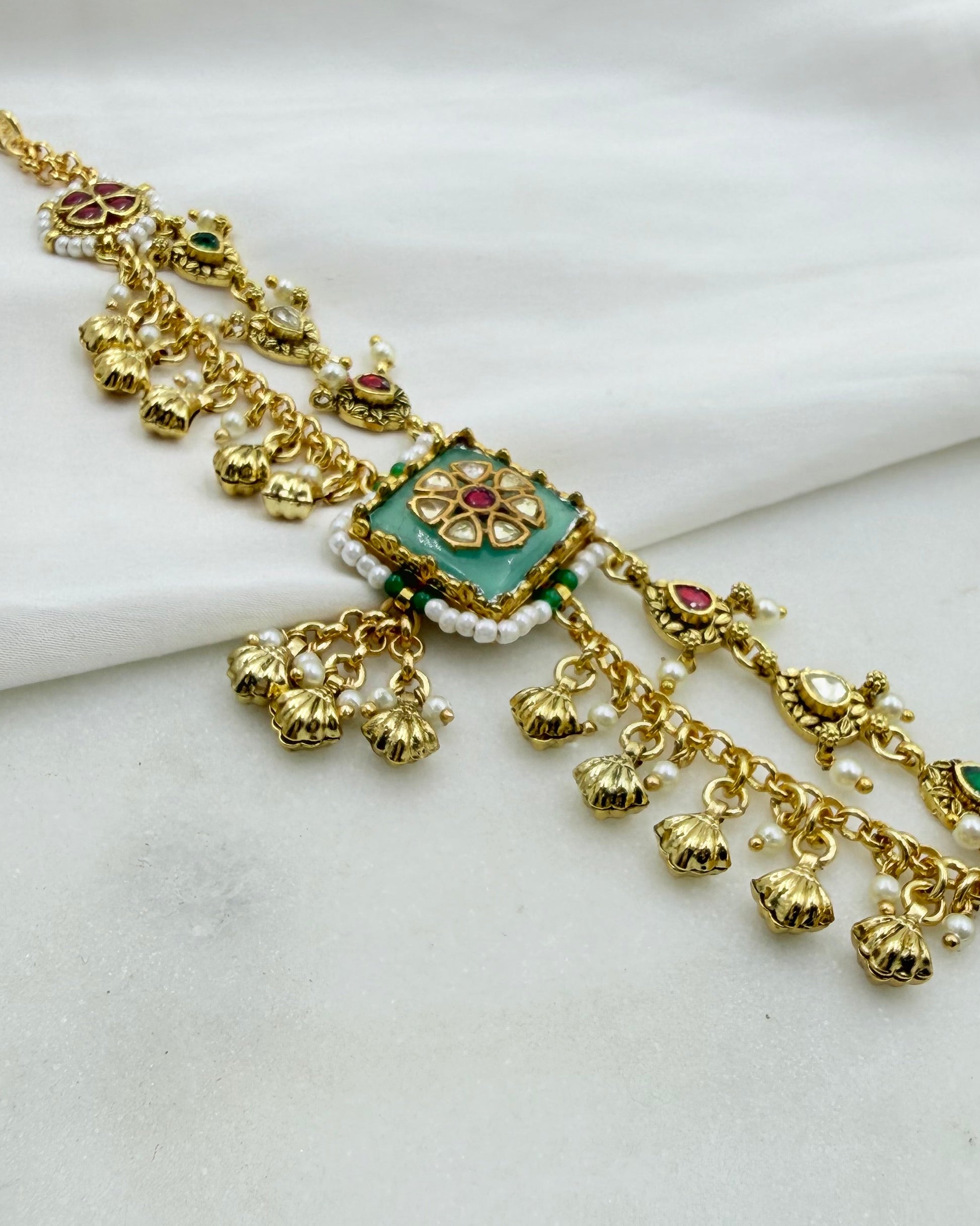 Gold-toned traditional Indian jewelry piece with a central turquoise green square, red and green stone accents, and dangling bell charms, displayed on a white fabric background.
