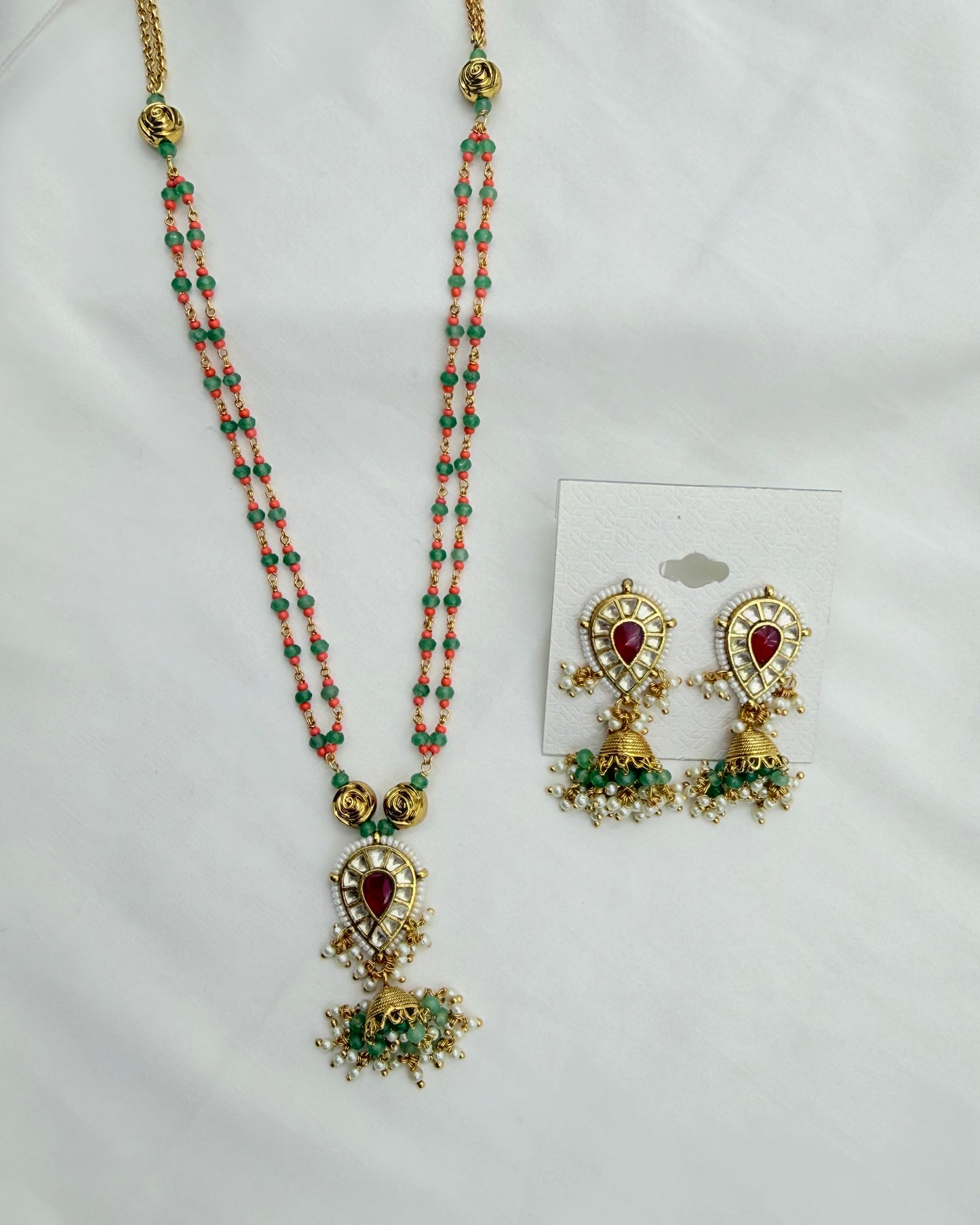 Gold-plated traditional Indian jewelry set featuring a long beaded necklace and matching earrings. The necklace has coral-pink and green beads with a central gold teardrop pendant containing a red heart-shaped stone and green accents.
