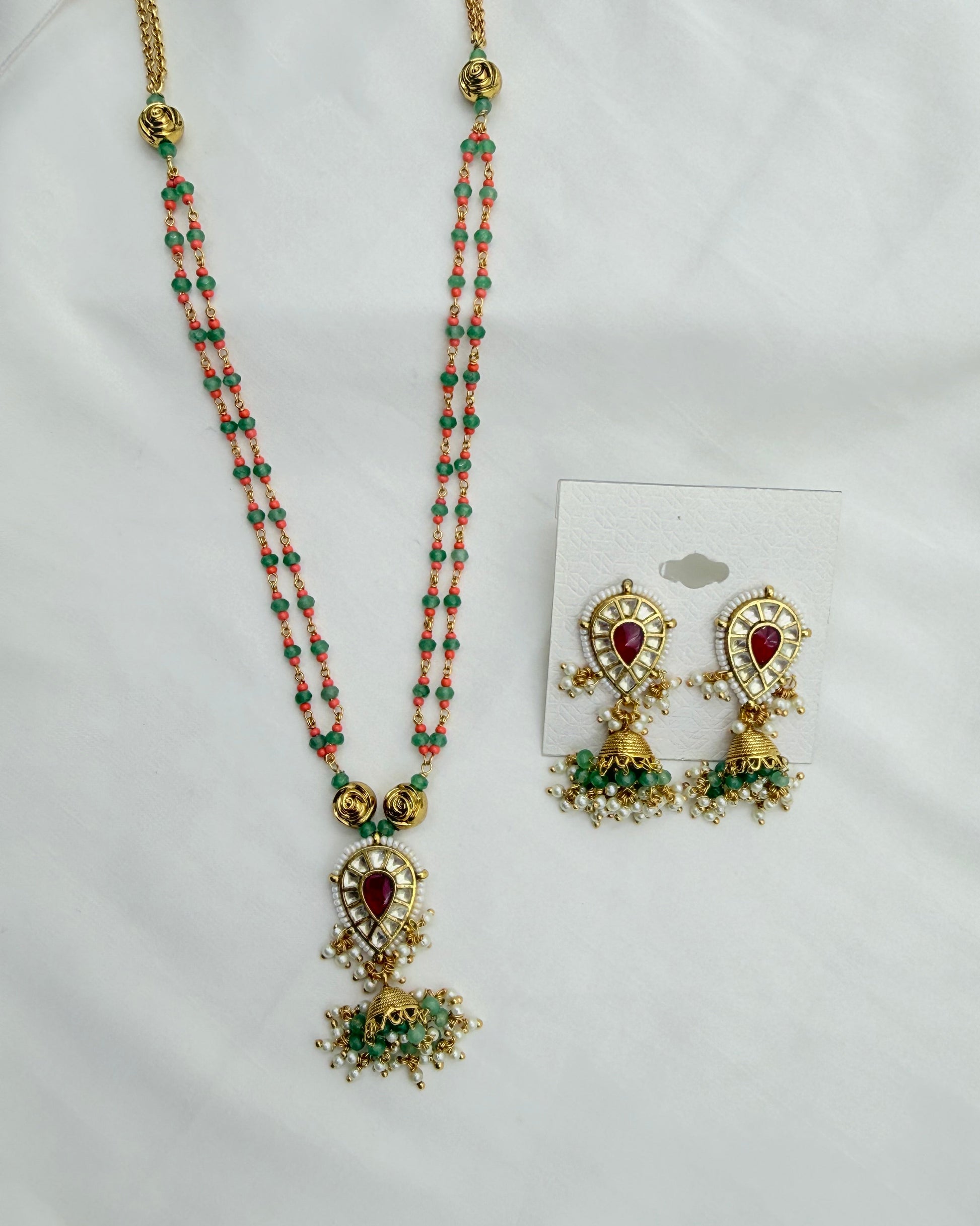 Gold-plated traditional Indian jewelry set featuring a long beaded necklace and matching earrings. The necklace has coral-pink and green beads with a central gold teardrop pendant containing a red heart-shaped stone and green accents.