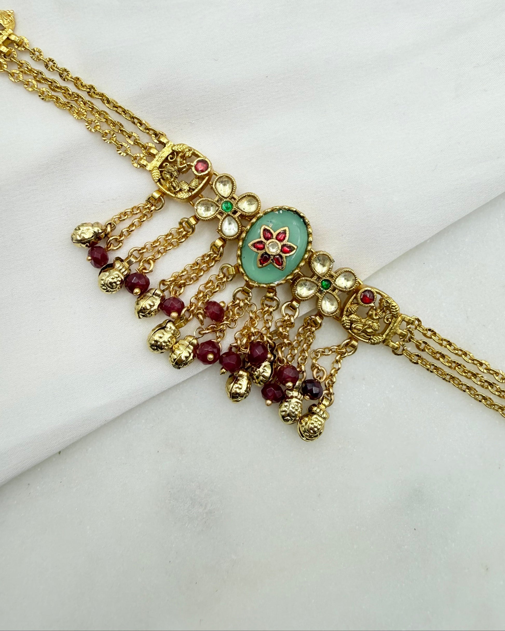 Gold-toned traditional Indian jewelry piece featuring a central oval light green stone with a red floral motif, surrounded by small red and green stones, with dangling beaded chains on a white marble background.