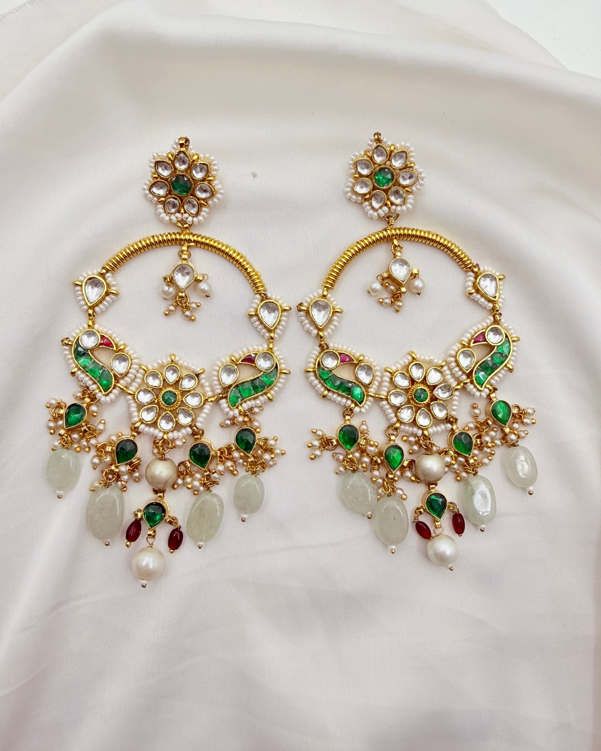 A pair of ornate gold-toned Indian chandbali earrings decorated with white faux pearls, green stones, and small red stones, displayed on a draped white fabric background.