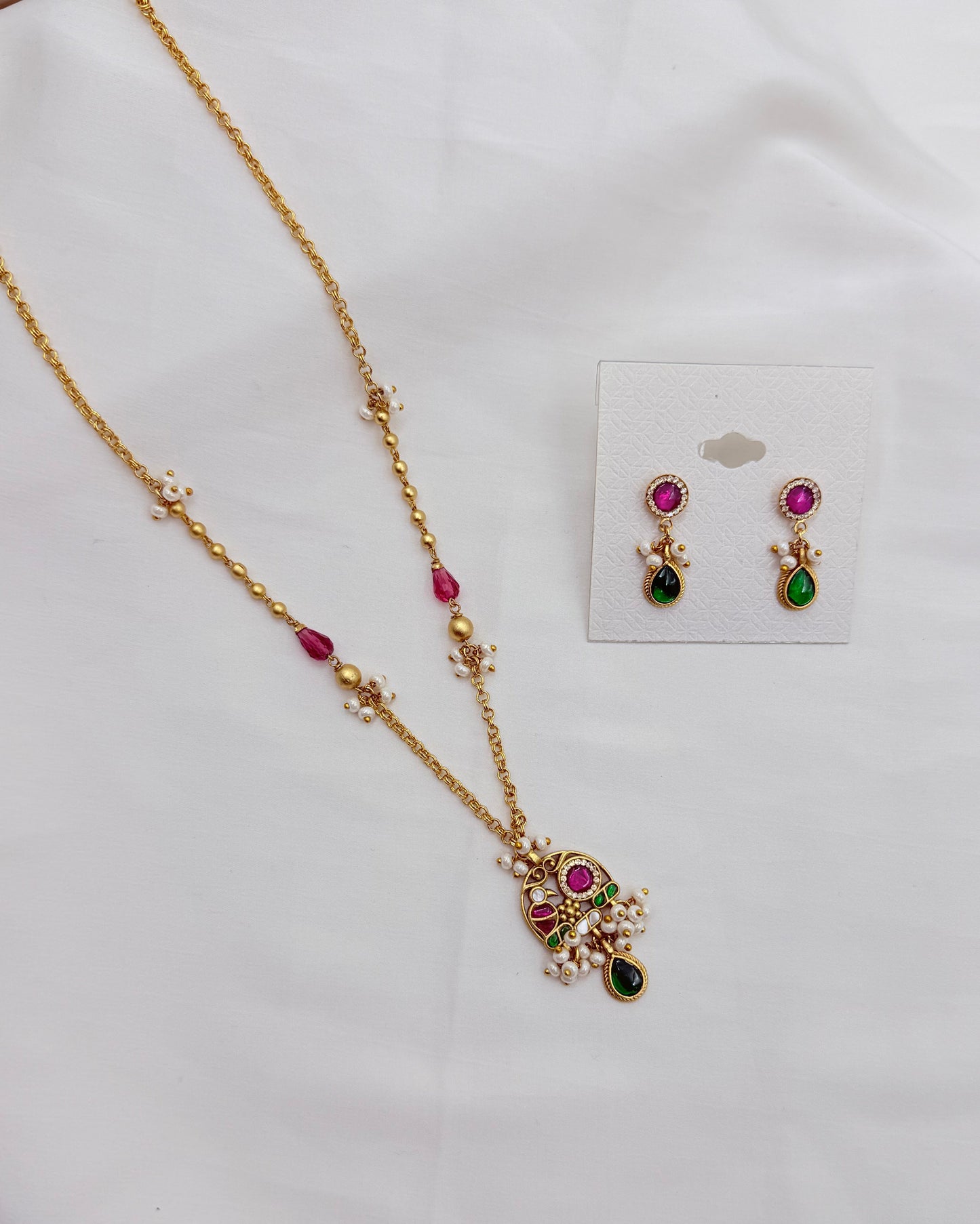 Gold-toned jewelry set with a necklace and matching earrings, decorated with pink, green, and white stones, displayed on a white fabric background with the earrings on a small card.