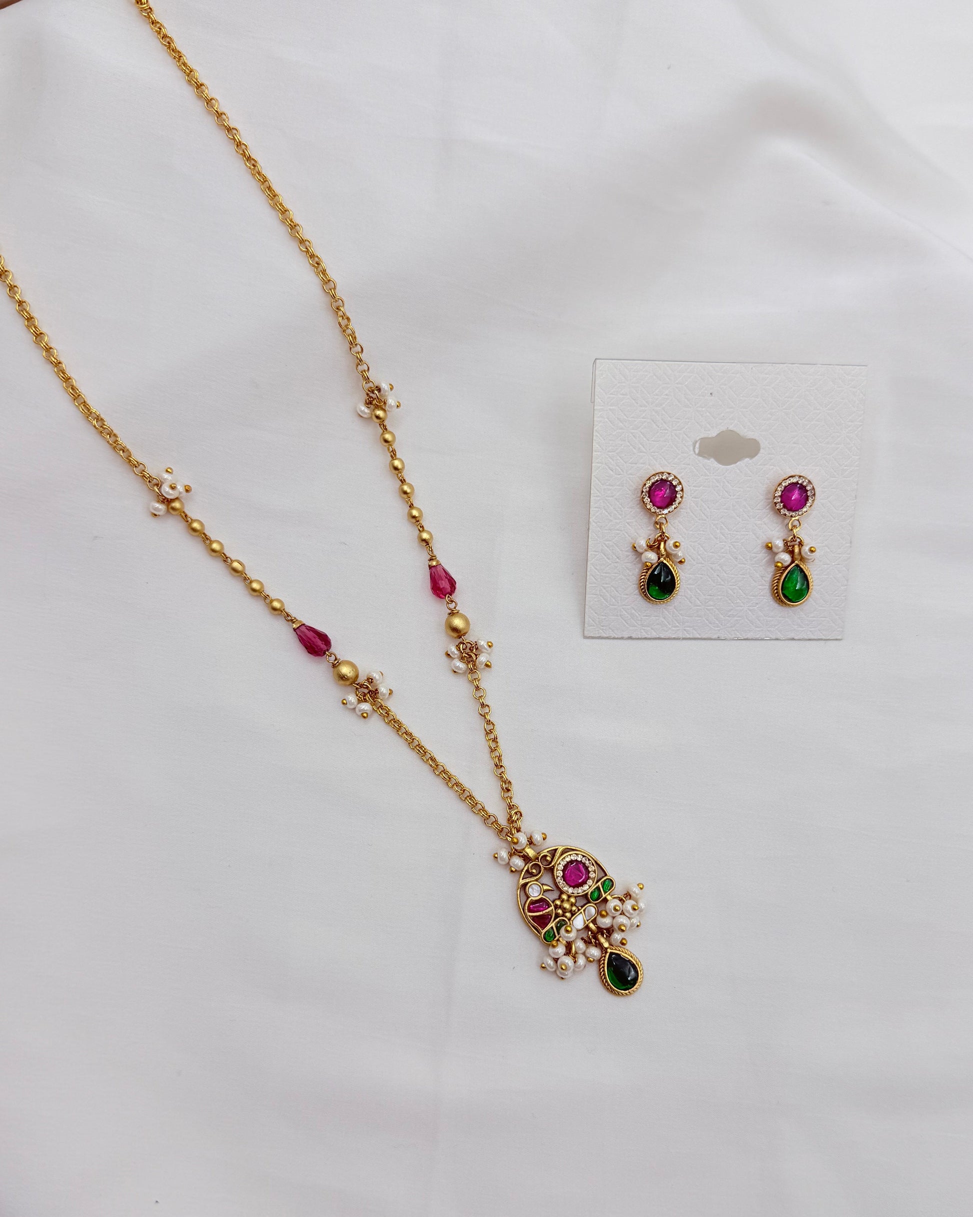 Gold-toned jewelry set with a necklace and matching earrings, decorated with pink, green, and white stones, displayed on a white fabric background with the earrings on a small card.